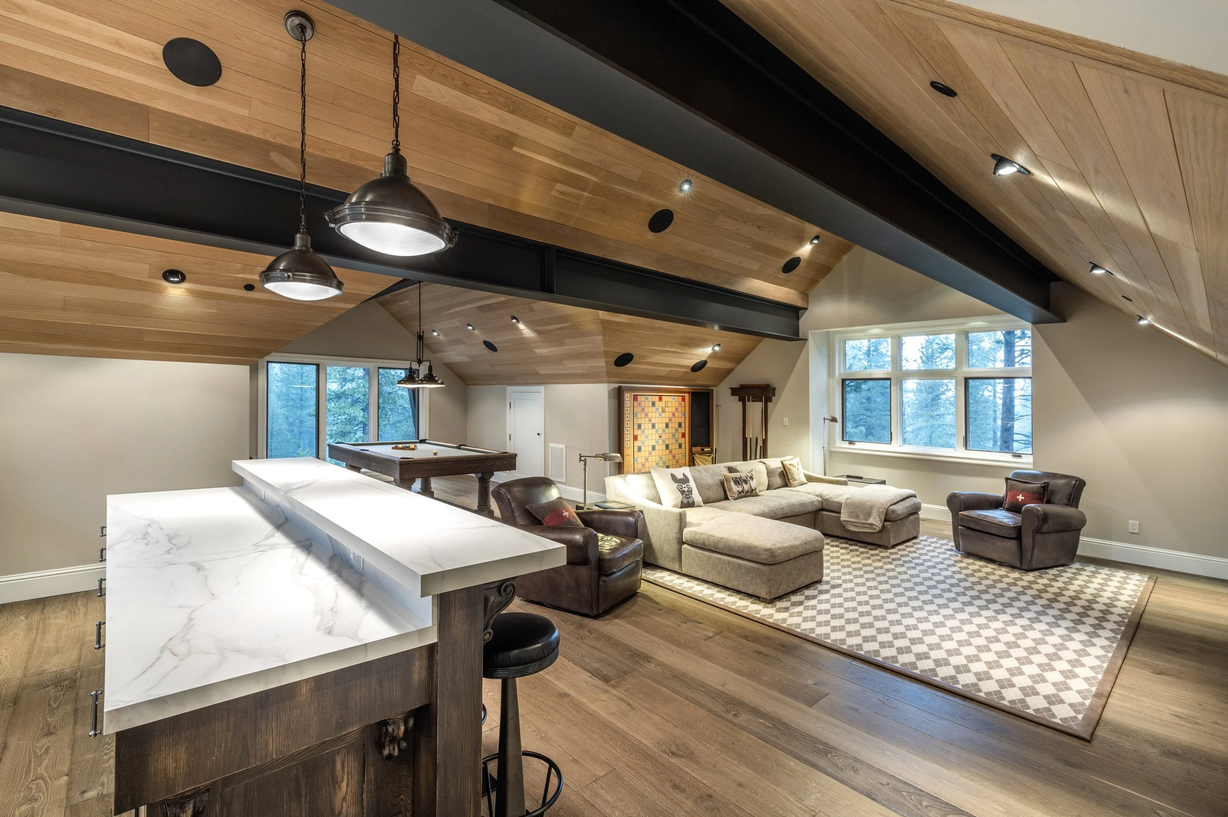 A cozy finished basement with wooden ceiling, large windows, a beige sectional sofa, two dark armchairs, a checkered rug, a pool table, and a marble kitchen counter in the foreground.