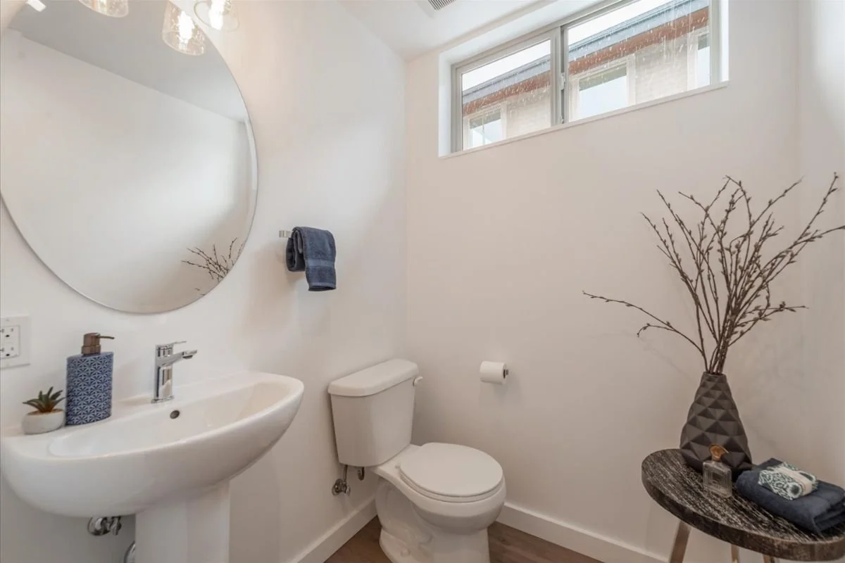 A small bathroom with a white sink, a toilet, a large round mirror, a window near the ceiling, a dark towel hanging on a hook, a small plant and decorative items on a side table, and a vase with dried branches.