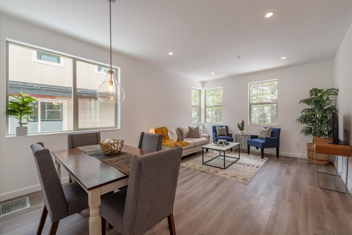 Open-concept living and dining area with large windows, a wooden dining table with gray upholstered chairs, a white sofa with throw pillows, two blue armchairs, a coffee table, and various potted plants.