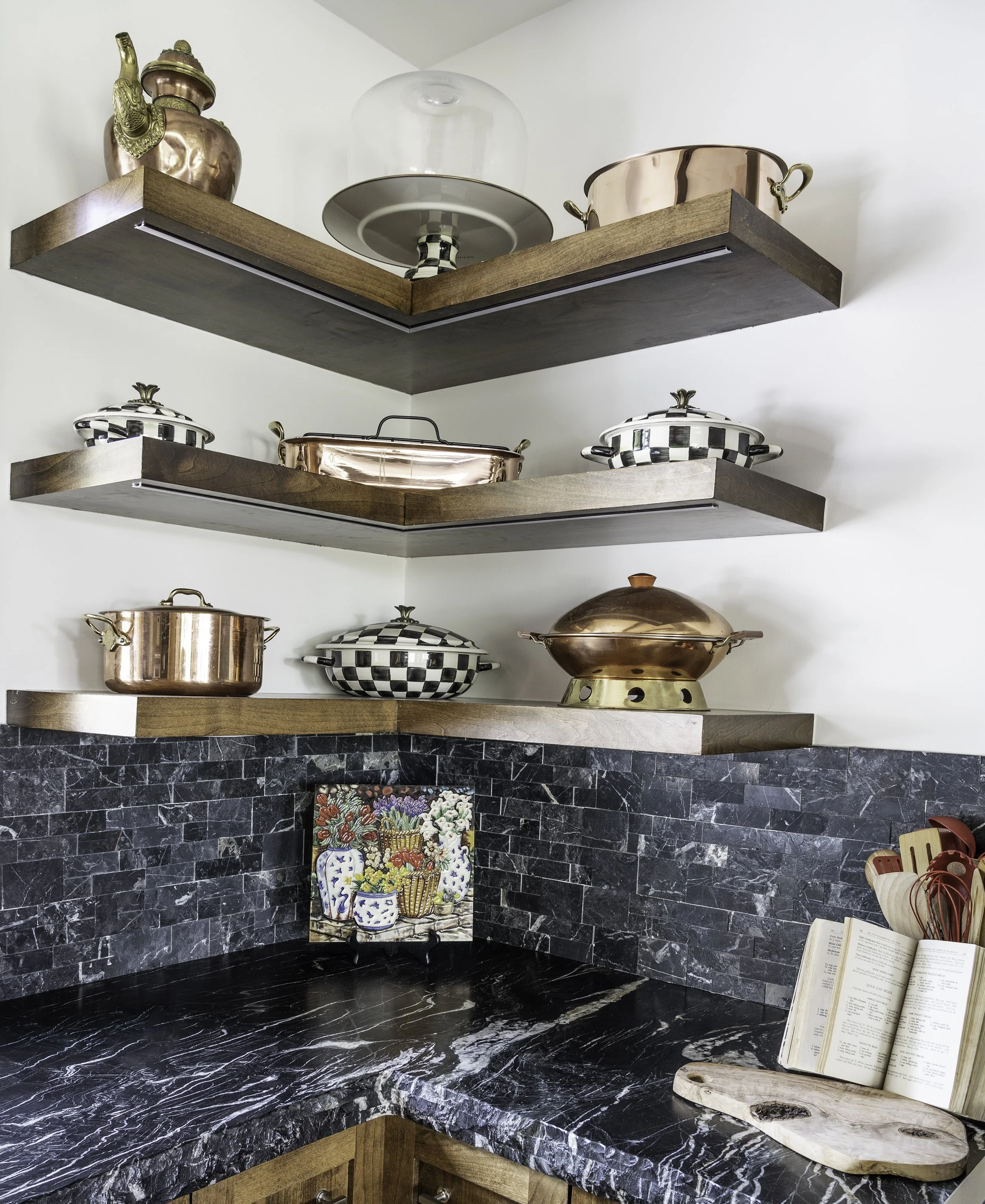 Kitchen corner with three wooden shelves holding copper and decorative cookware and containers, black marble countertop with a colorful floral painting, open cookbook, and red utensils in a container.