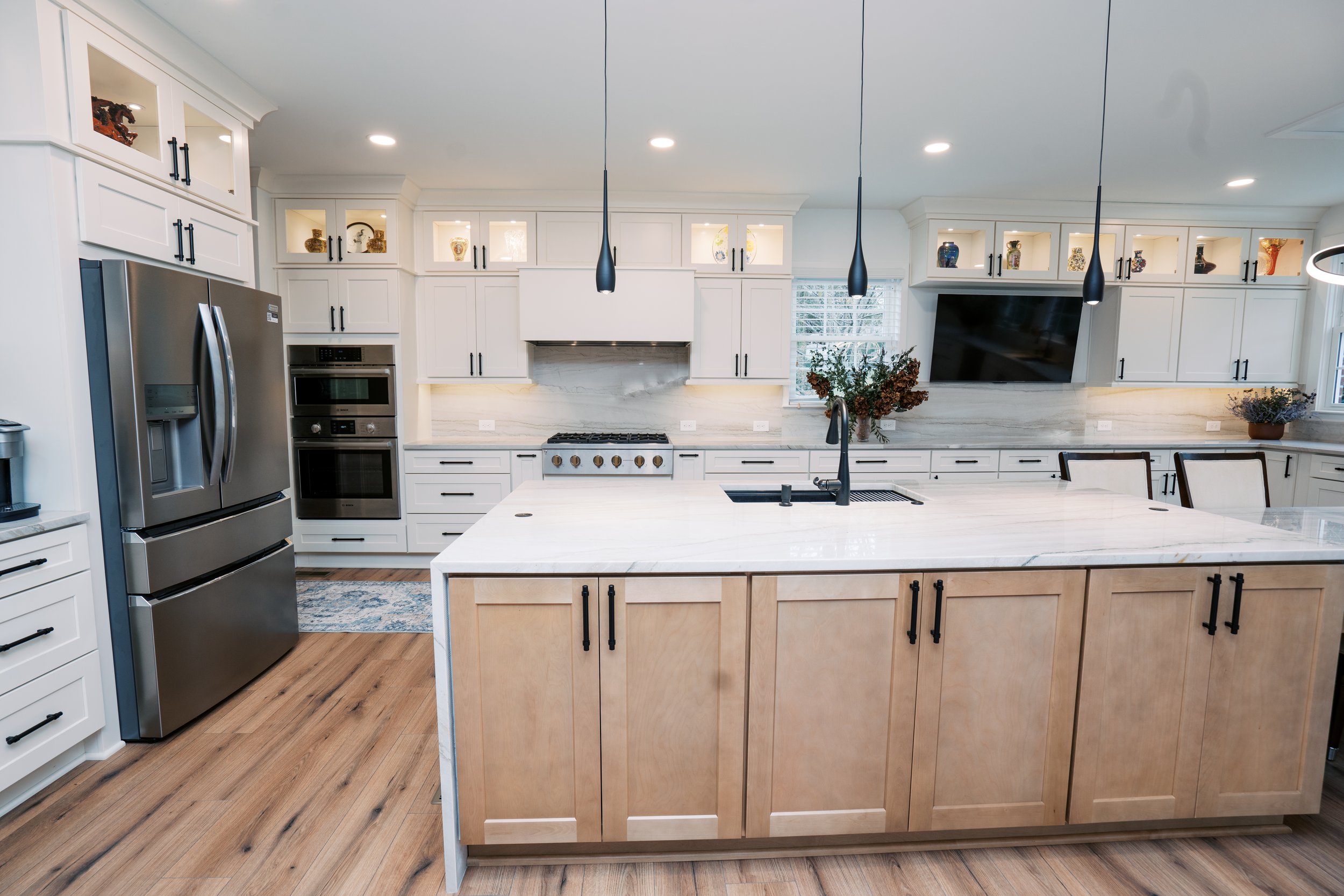 Modern kitchen with white cabinets, stainless steel refrigerator, built-in oven, and a large kitchen island with a white marble countertop, black hardware, pendant lights, and a black sink with a black faucet.