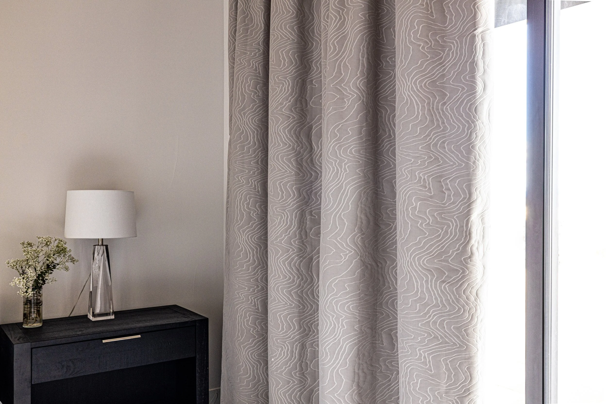 A corner of a room with a black cabinet, a white table lamp with a clear base, and a small vase with white flowers on top, next to a patterned curtain.