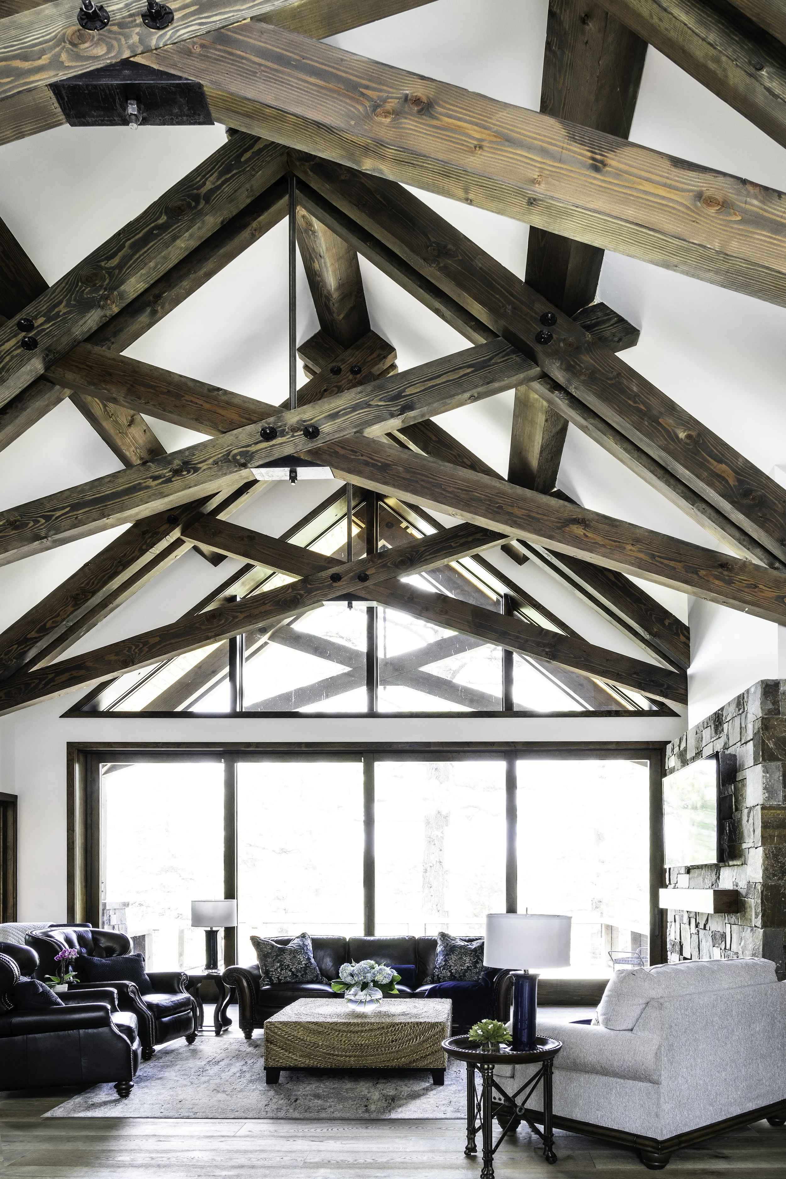 Living room with high vaulted ceiling featuring exposed wooden beams, large windowed wall, stone fireplace, and various seating including leather and fabric sofas and armchair, coffee table with flowers, and side tables with lamps.