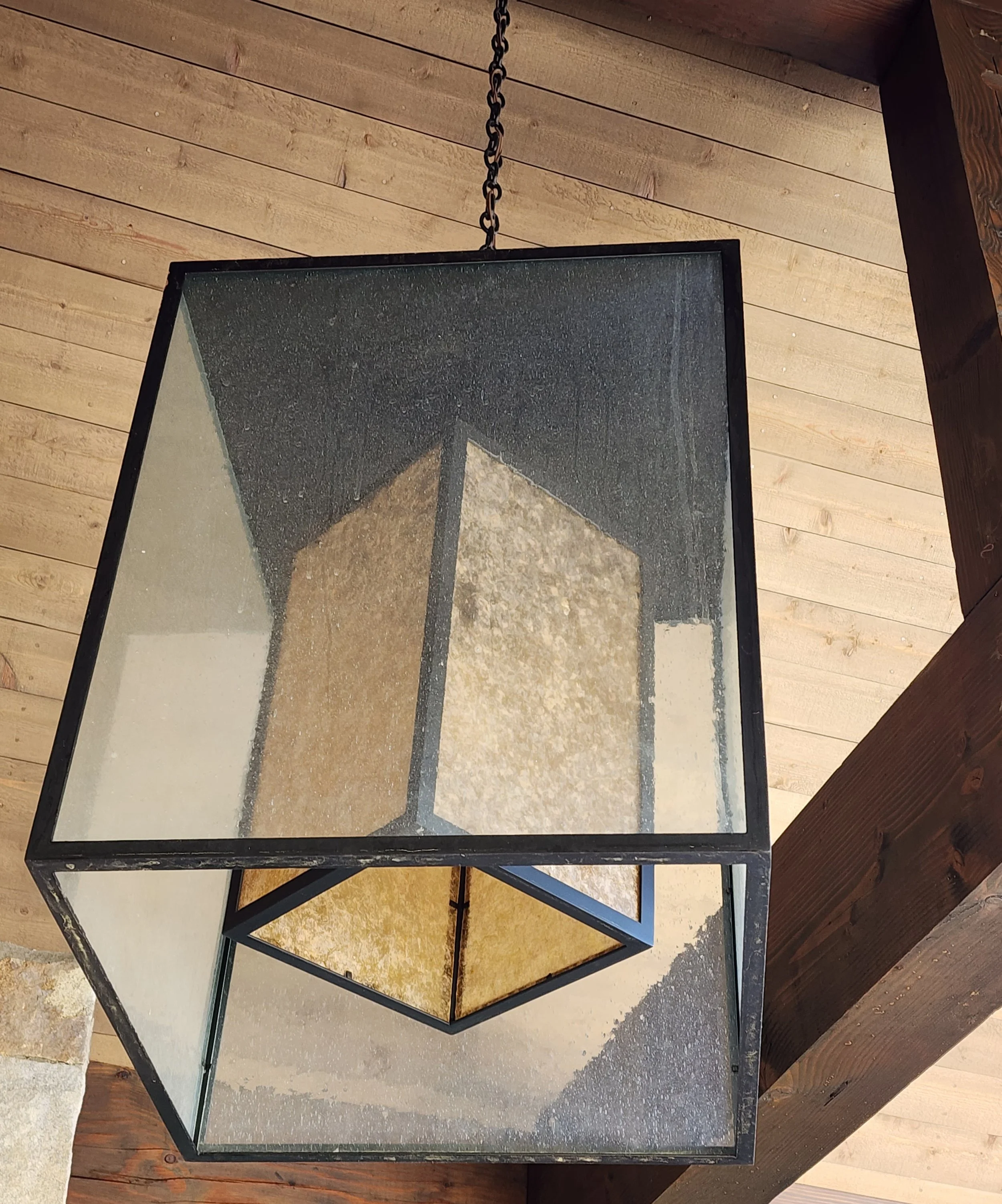 A modern geometric pendant light fixture hanging from a wooden ceiling, with a combination of black metal frame and glass panels on the outside and a mica and black framed interior shade. Custom designed by Emily Roose.