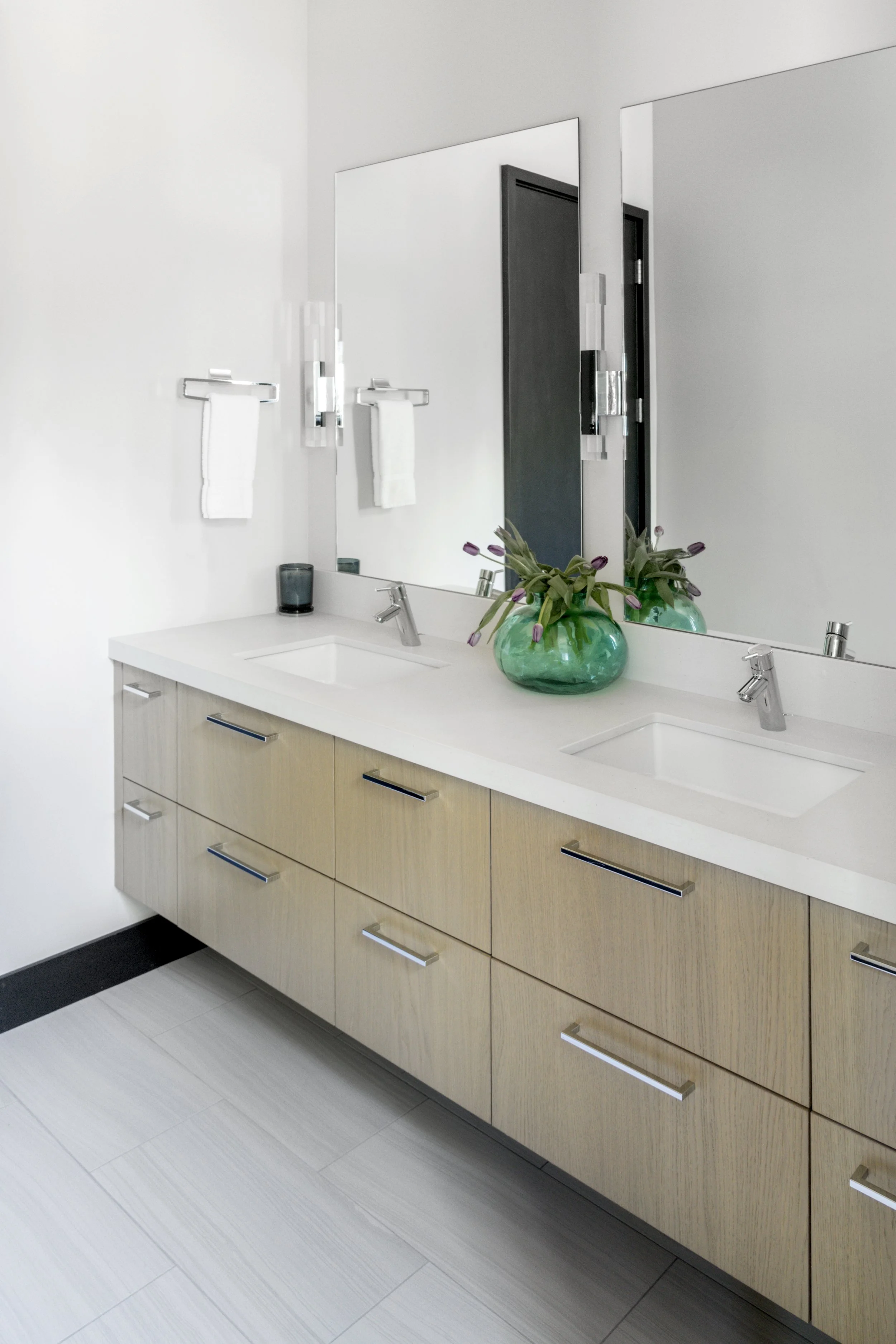 Modern bathroom vanity with dual sinks, large mirror, white countertop, beige drawers with sleek handles, a green vase with purple flowers, and wall-mounted light fixtures.