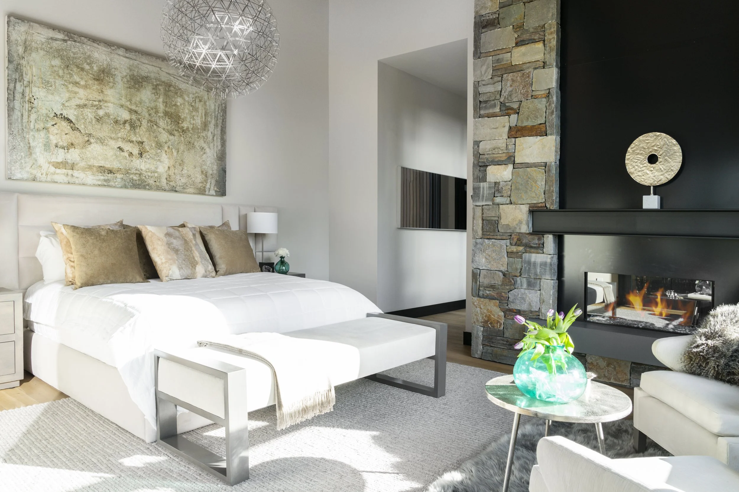 A modern bedroom with a white bed, beige and gold pillows, a painting above the bed, a side table with a lamp and a vase, a fireplace with a decorative sculpture above it, a stones wall, a green glass vase with flowers on a small table, and a white a