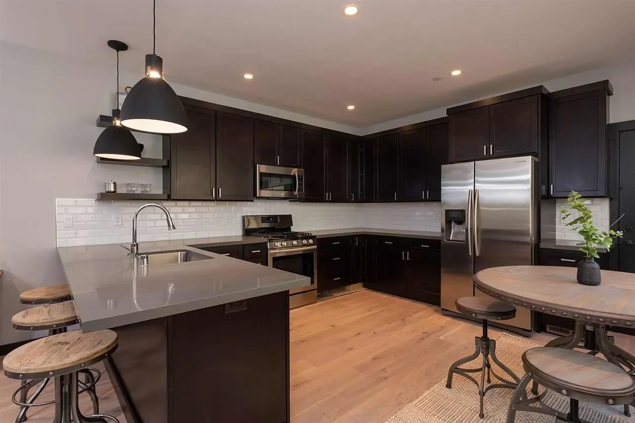 Modern kitchen with black cabinets, stainless steel appliances, white subway tile backsplash, and a kitchen island with a sink and three wooden stools. There is a dining table with a plant and a black vase, and black pendant lights hanging from the c