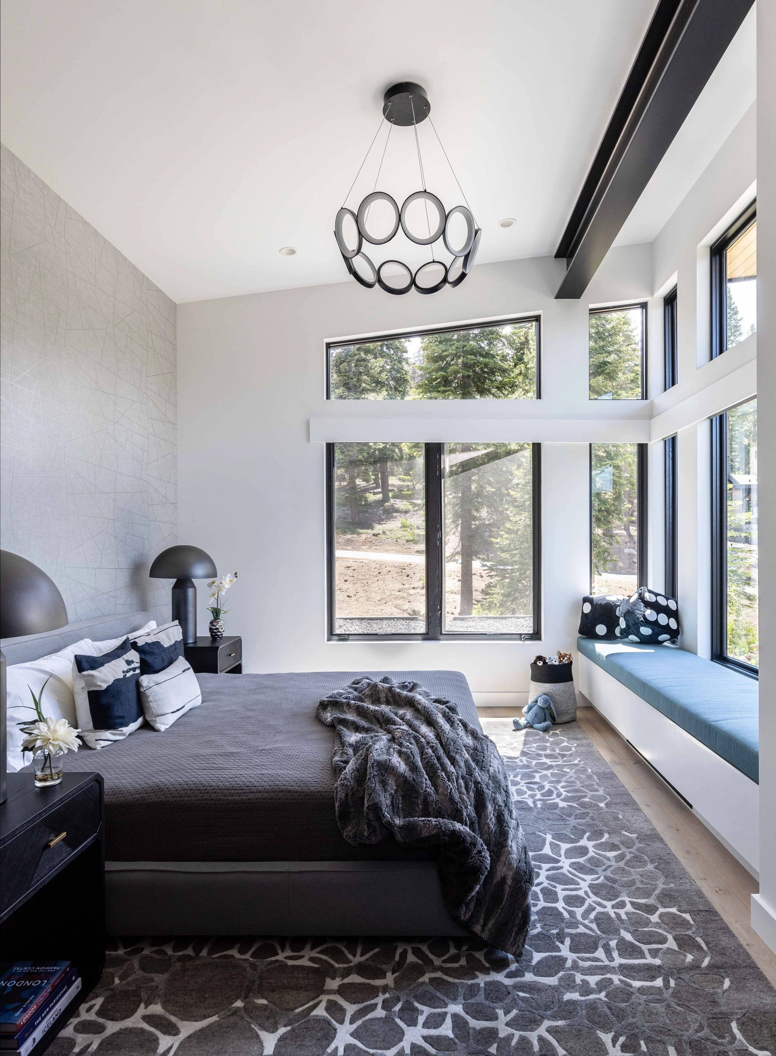 Modern bedroom with large windows, a bed with a dark grey bedspread and striped pillows, a black nightstand with a lamp and a flower in a vase, a patterned rug, a window seat with cushions, and a ceiling light fixture.