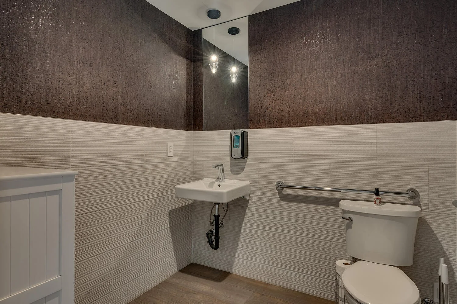 Accessible bathroom with brown textured upper walls, beige lower tiled walls, a wall-mounted sink, a toilet with a grab bar, and a mirror with hanging lights.