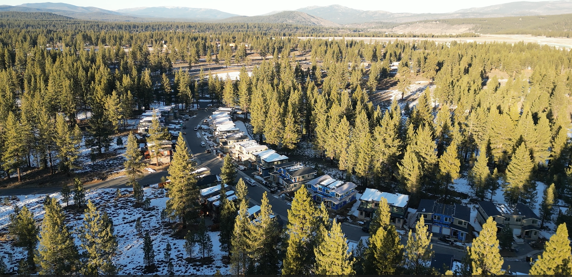 Aerial view of a mountain residential area with rows of modern houses surrounded by pine trees, some with snow on rooftops, during daytime.