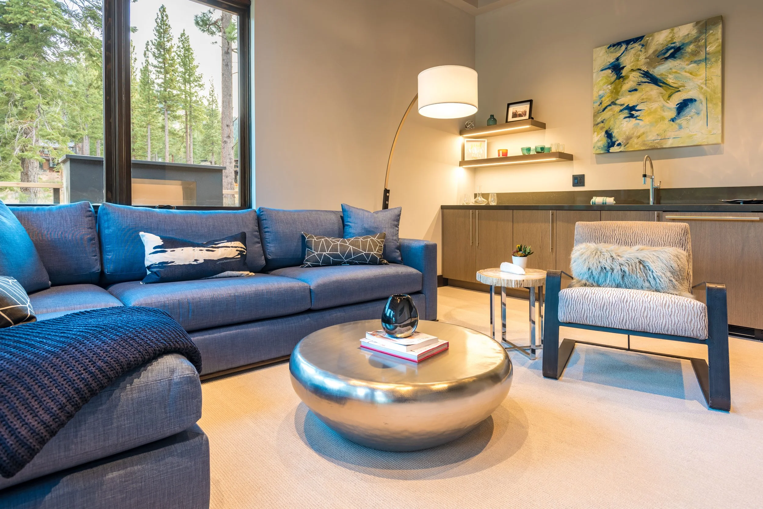 Living room with blue sofa, abstract painting, armchair with fur pillow, round coffee table, tall floor lamp, and open kitchen with wooden cabinets, shelves, and natural light from large window showing trees outside.