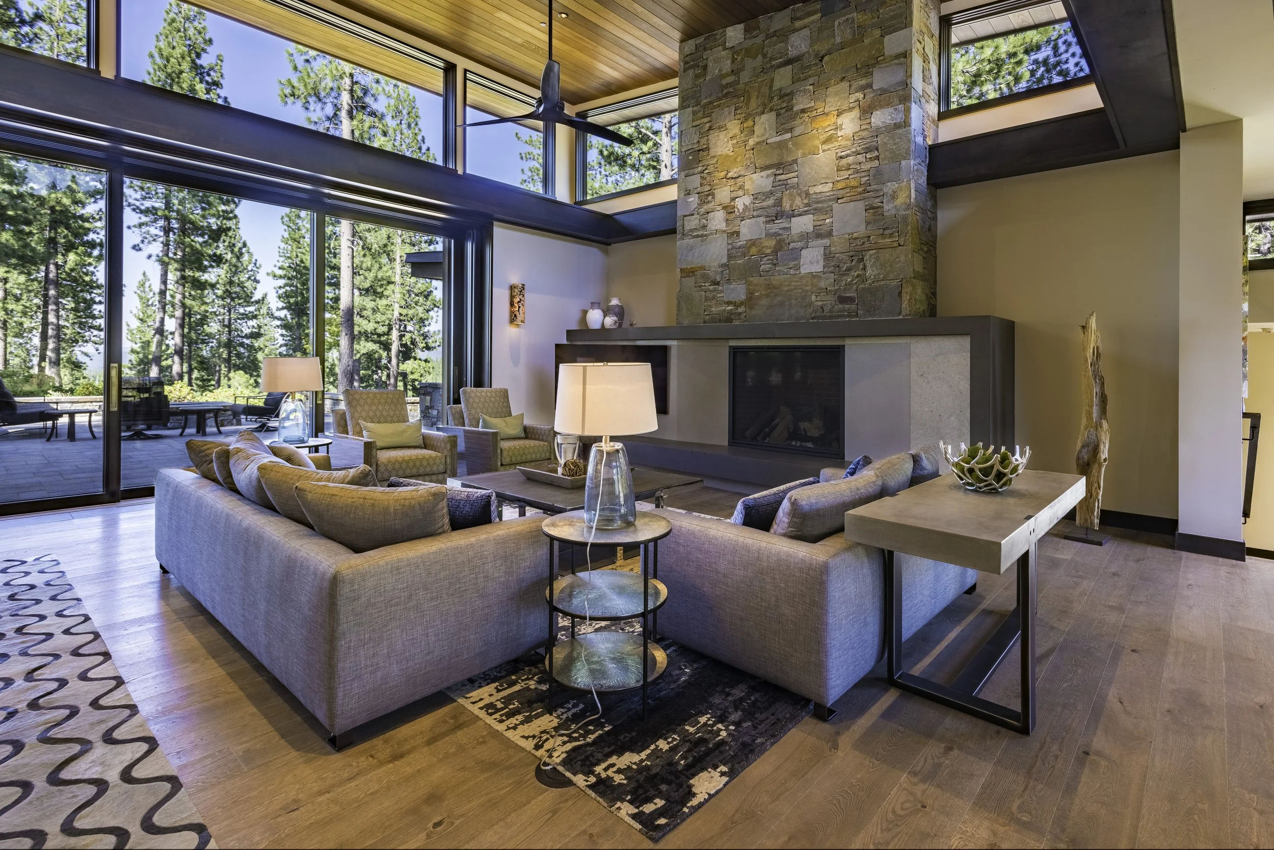 Living room with large glass windows, wooden ceiling, stone fireplace, modern furniture, and scenic forest view.