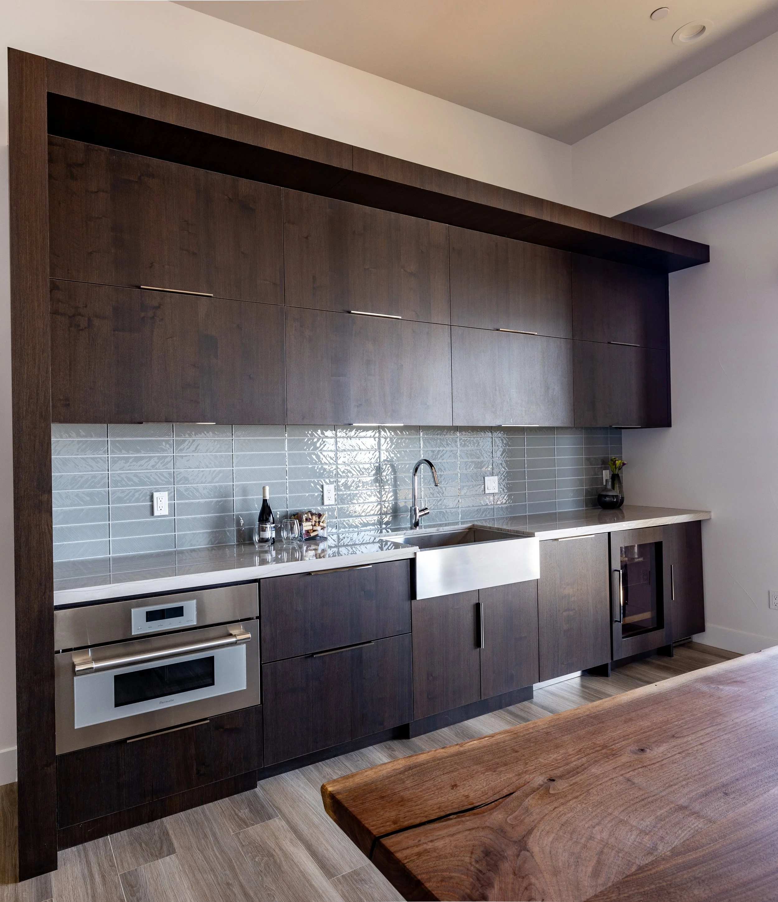 Modern kitchen with dark wood cabinetry, a stainless steel sink, backsplash tiles, built-in oven, and small appliances on the countertop.