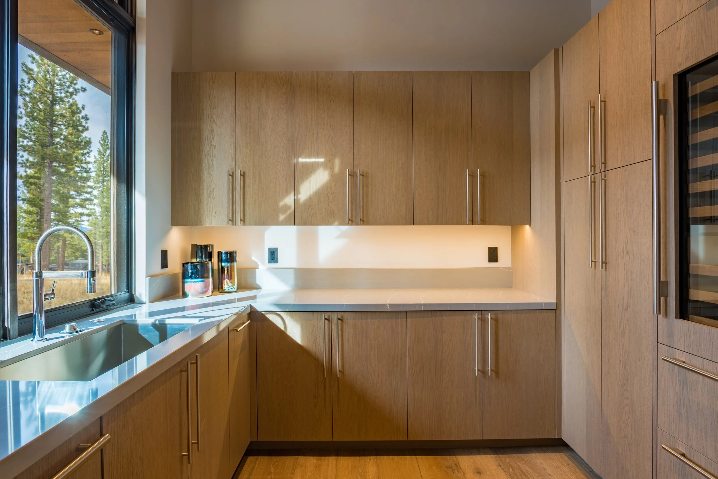 Modern kitchen with wooden cabinets, a large window with a view of trees, and a light-colored countertop with three decorative jars near the sink.