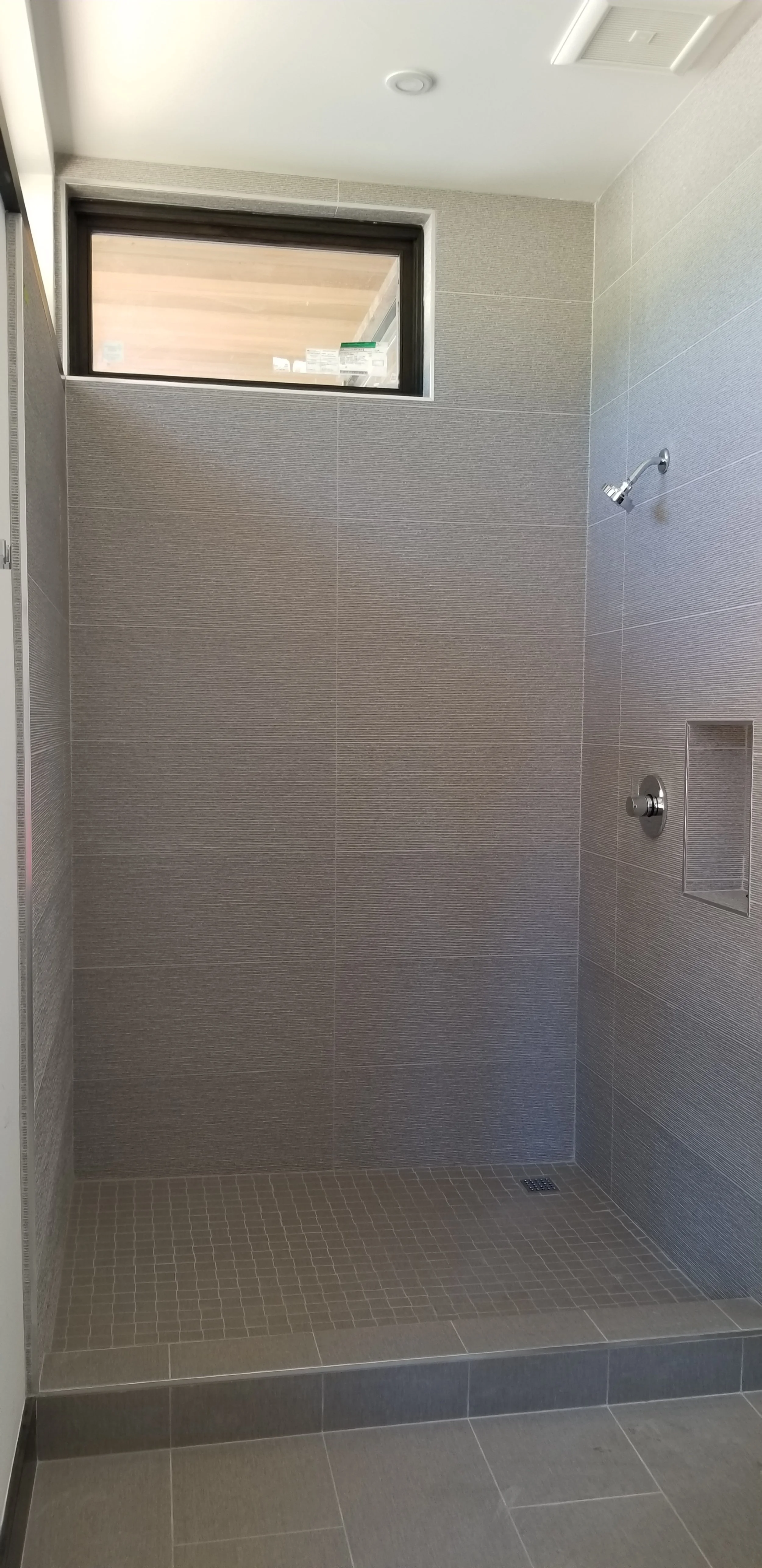 A modern, empty shower area with gray tiled walls and floor, a small window near the ceiling, and a wall-mounted showerhead.