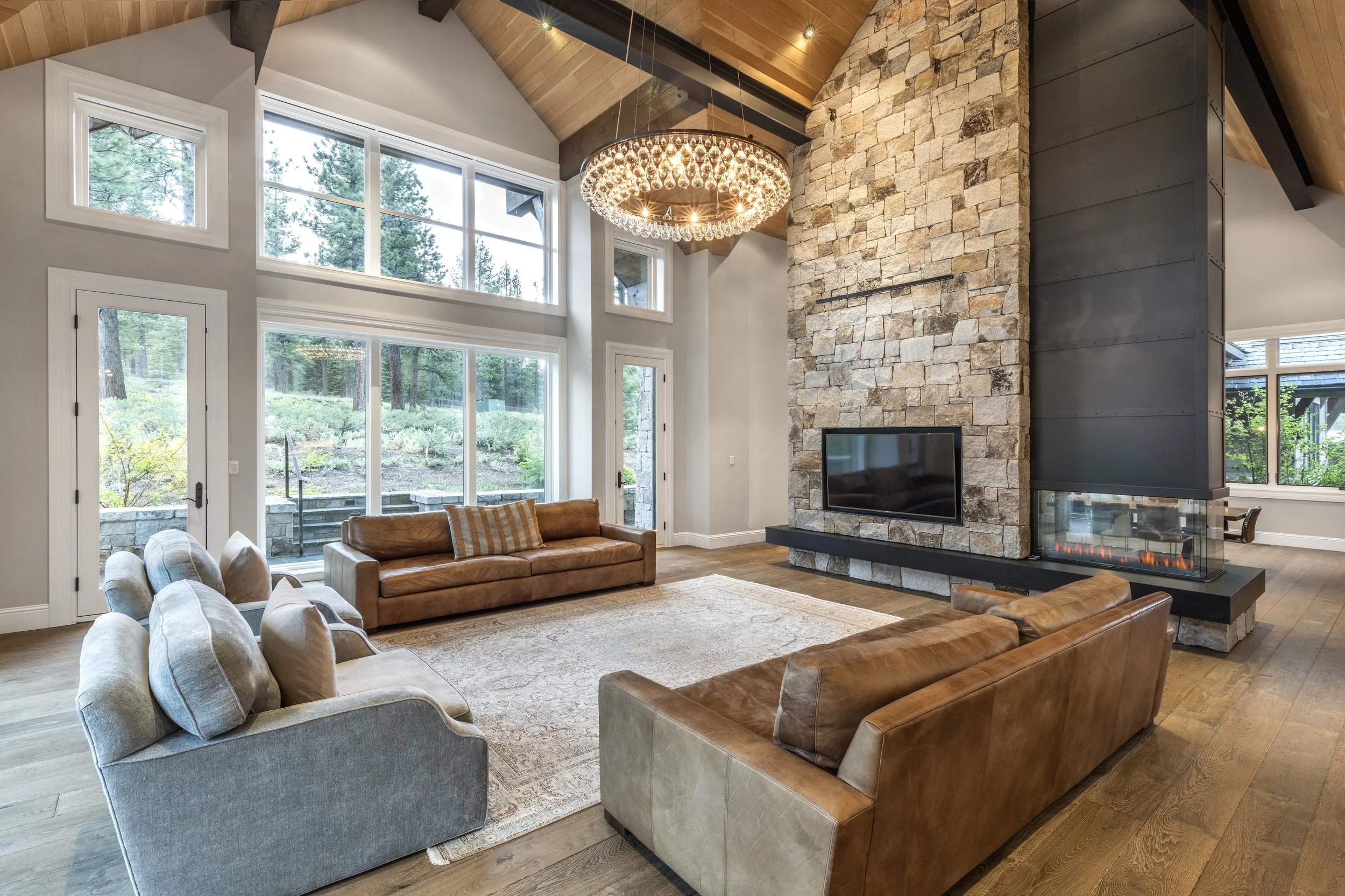 Living room with large windows, stone fireplace, and leather sofas, featuring a modern chandelier and wooden ceiling.