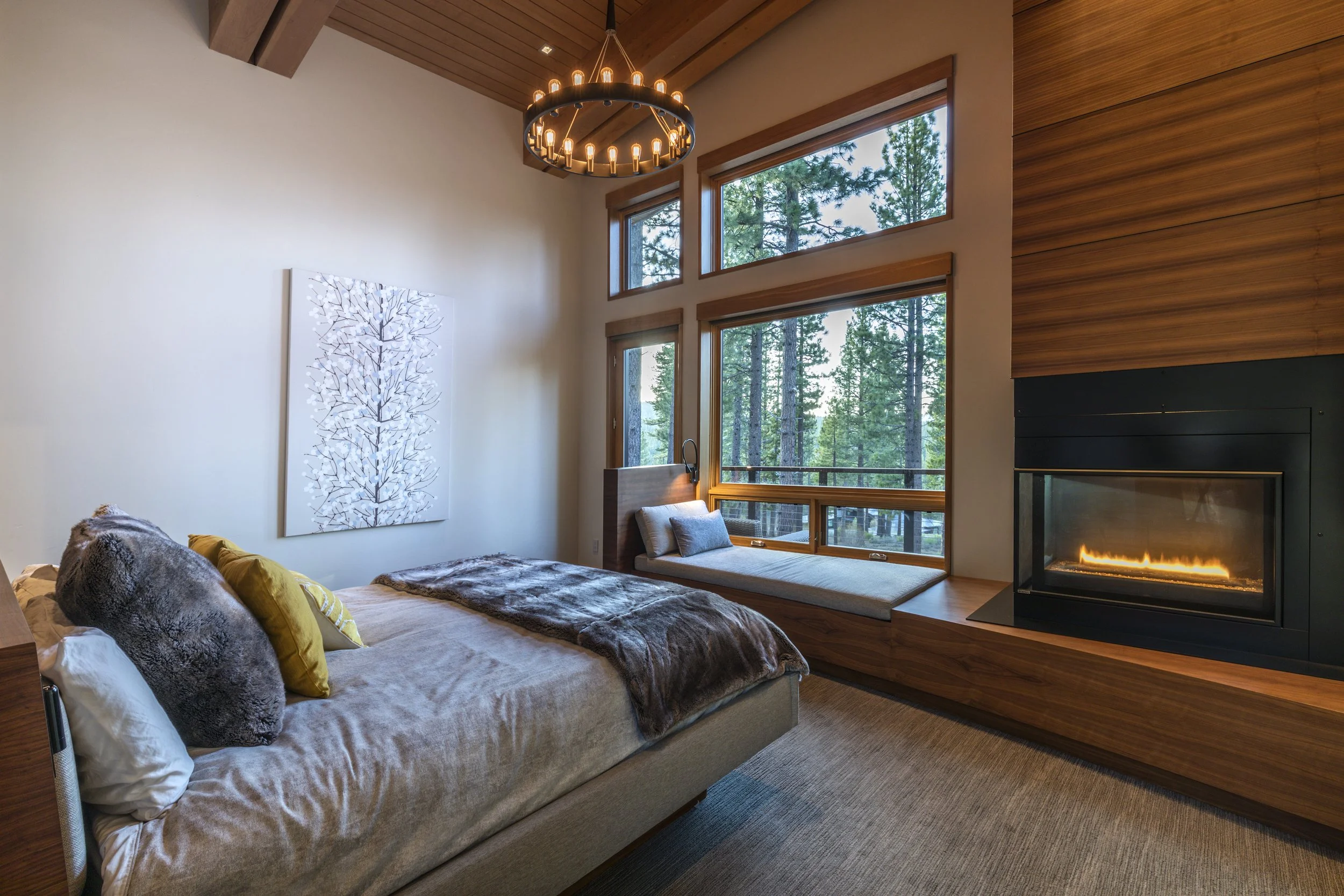 Cozy bedroom with large windows showing a forest view, a modern fireplace, a bed with fur and yellow pillows, a wall art of a tree, and a chandelier.