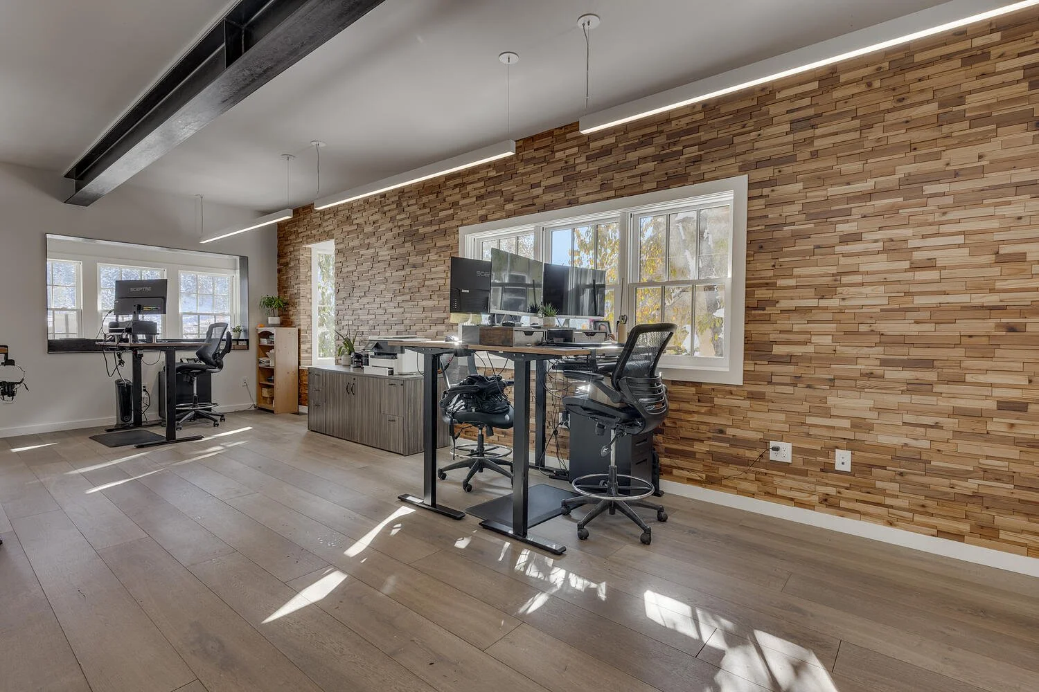 Modern office with wooden accent wall, large window, and two workstations with multiple monitors and office chairs.