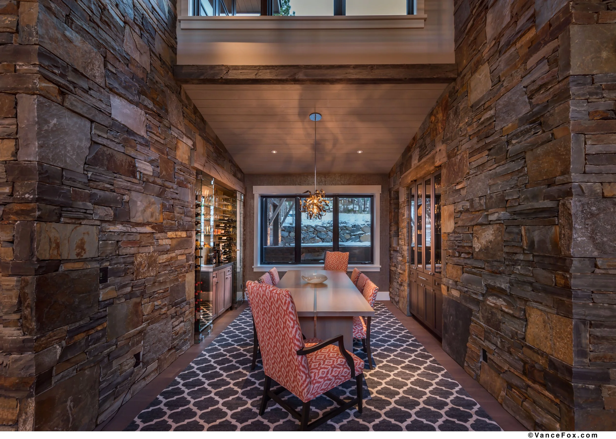 Interior of a dining room with stone walls, a custom dining table designed by Emily Roose, and chairs with patterned upholstery around the table. A modern chandelier hangs above the table, and built-in cabinets flank the room. 