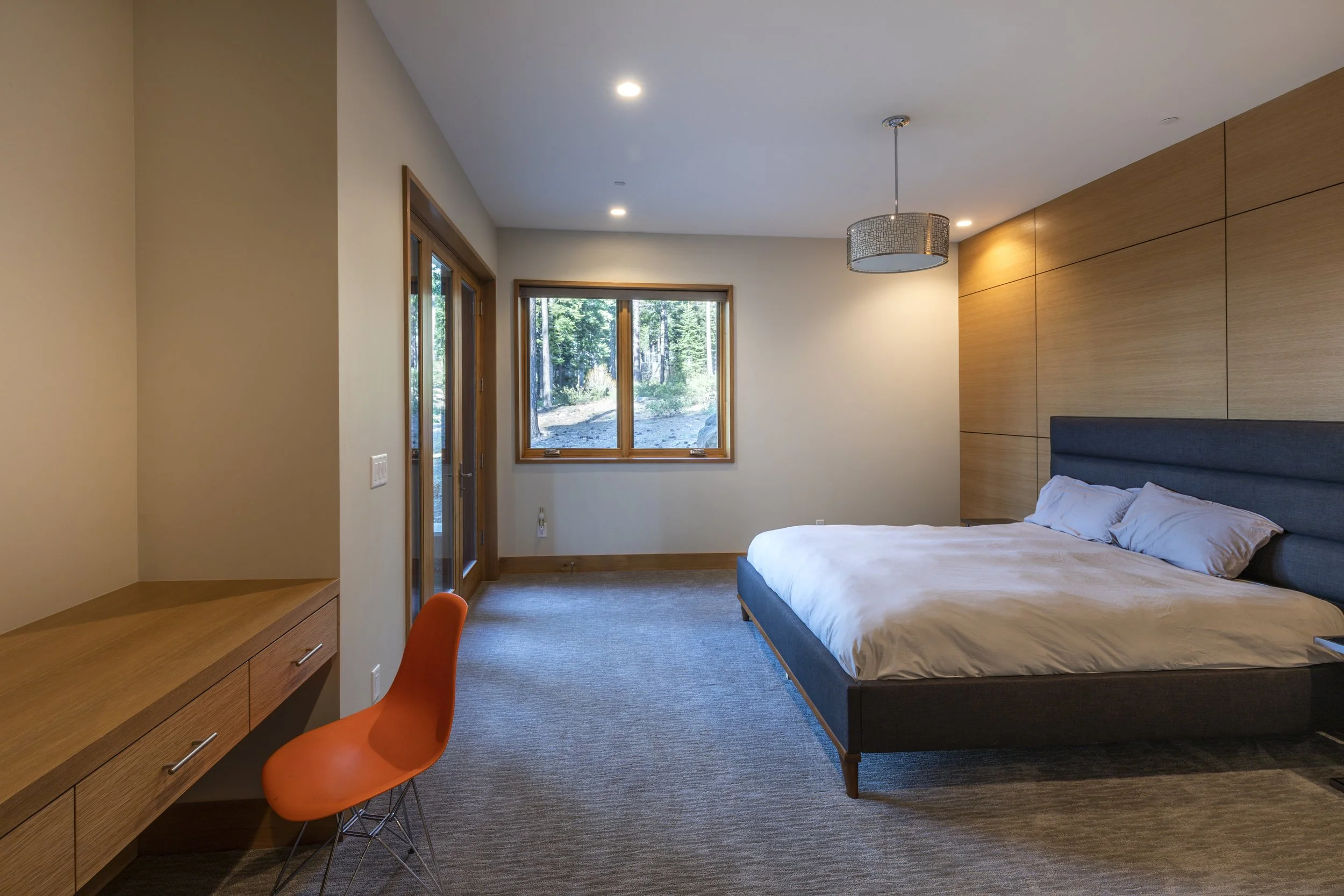 A modern bedroom with a large bed, wooden wall paneling, a window, a sliding glass door, a desk with an orange chair, a ceiling light, and a view of trees outside.