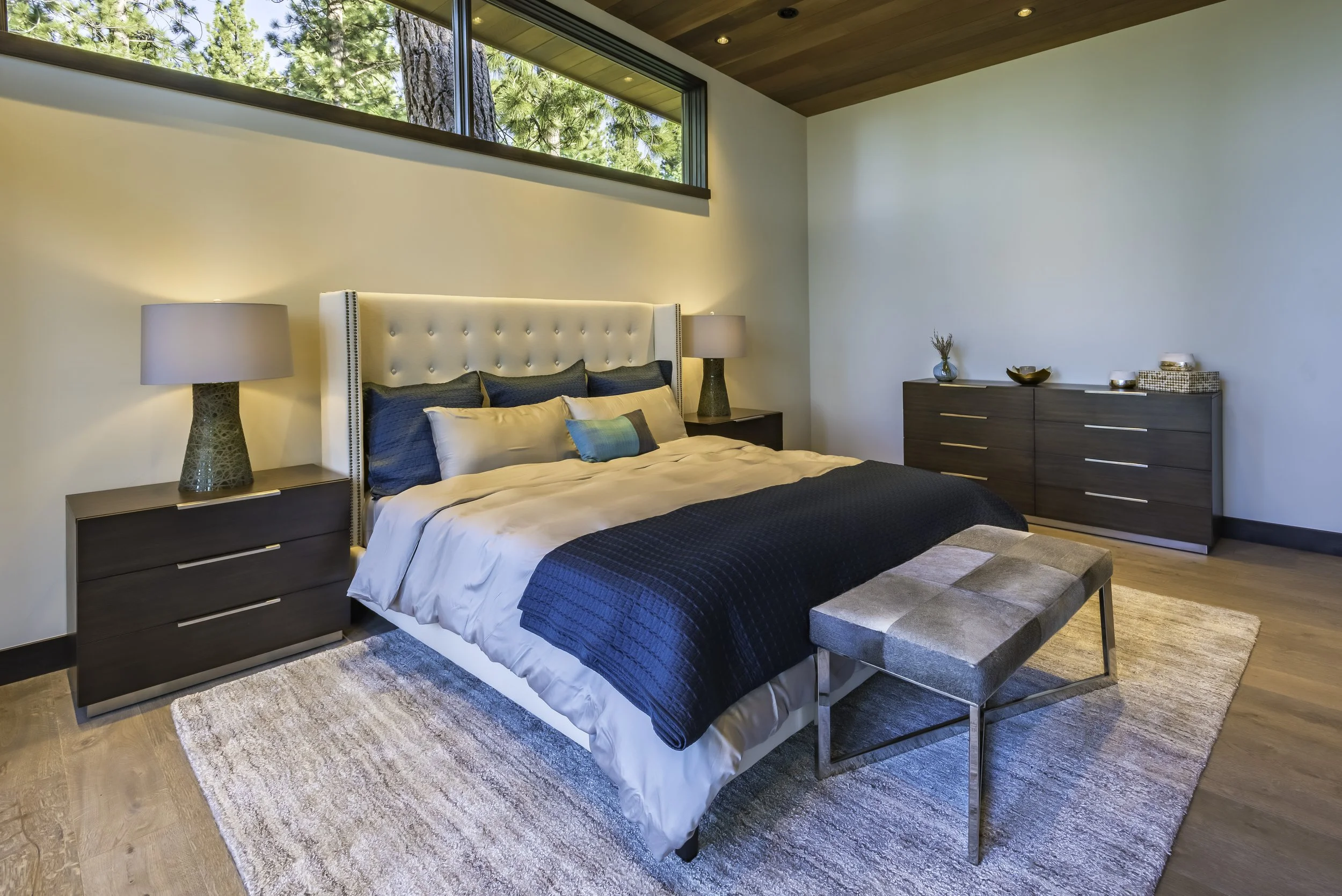 A modern bedroom with a large bed featuring a tall, tufted white headboard, navy and beige pillows, and a navy bedspread. There are two nightstands with lamps on each side of the bed, a beige area rug on wooden flooring, and a dark wood dresser with 