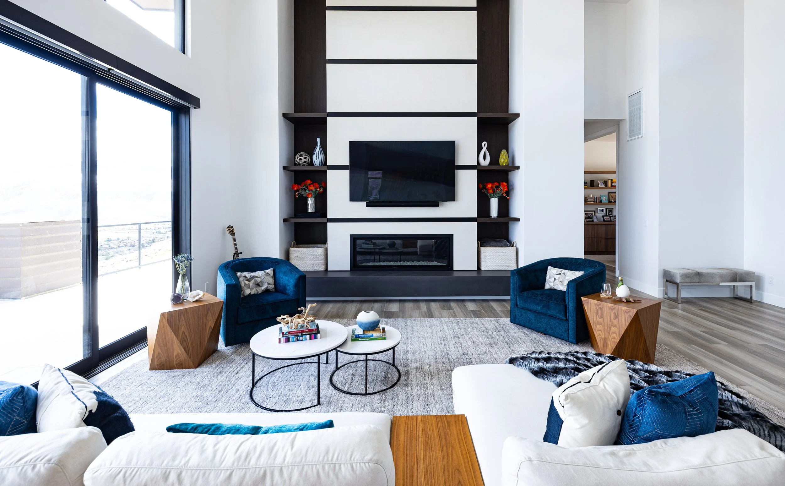 Modern living room with a white sofa, blue armchairs, a fireplace, a large flat-screen TV, and large windows with a view outside.