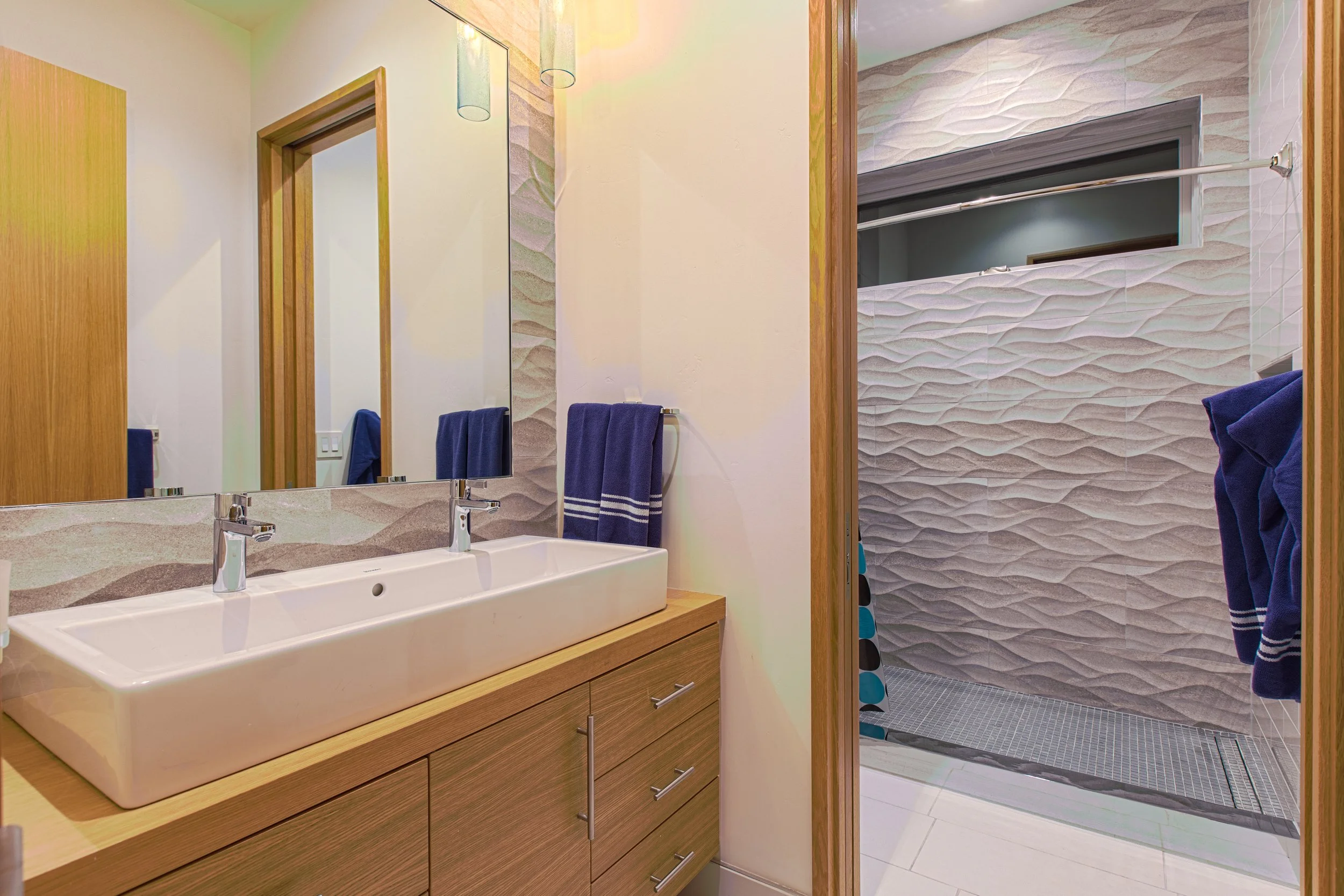 Modern bathroom with a rectangular vessel sink, wooden vanity, large mirror, and walk-in shower with wave-textured wall tiles and a glass window.
