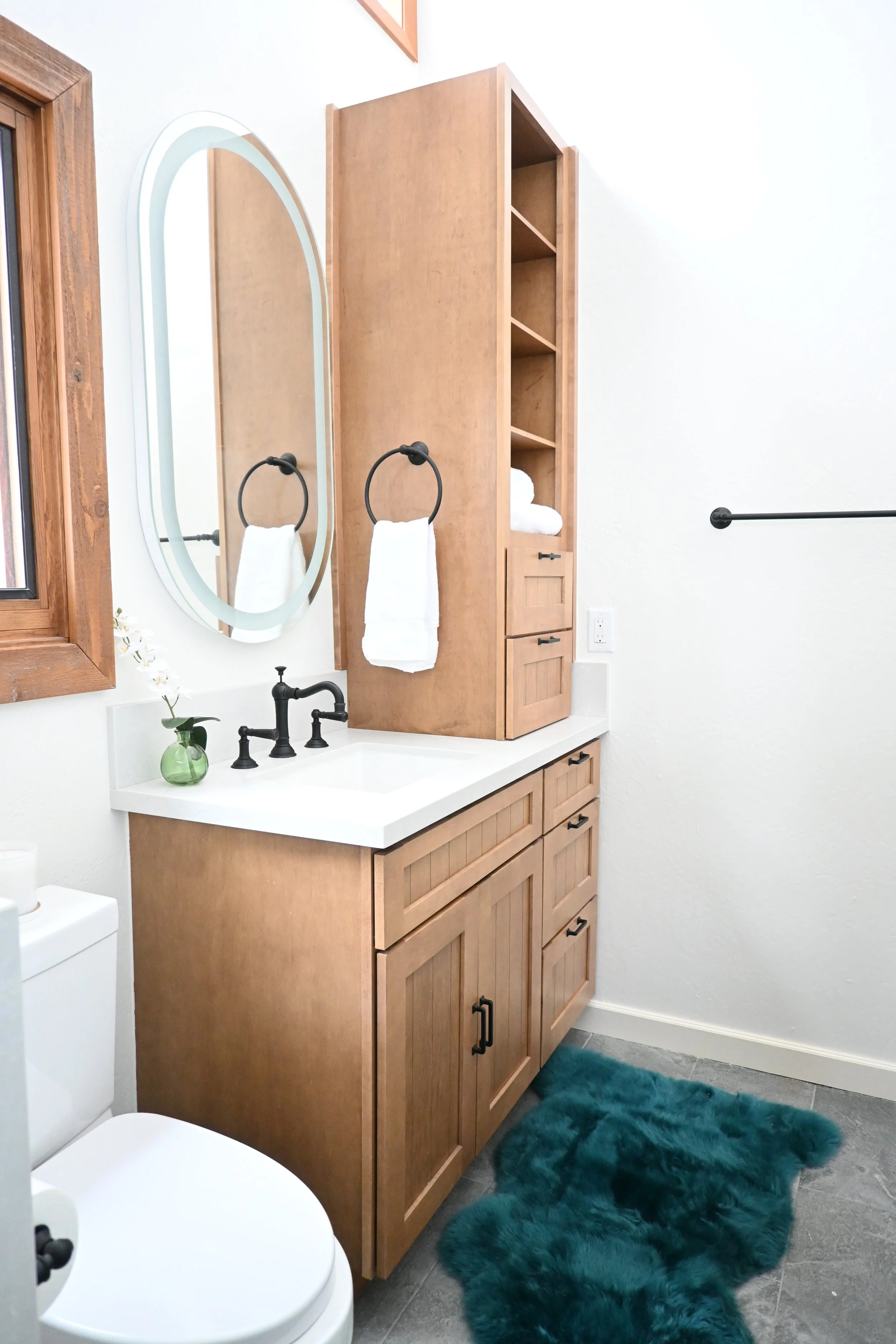 Bathroom with wooden cabinet, oval mirror, black faucet, white countertop, rolled white towels, small green vase with white flowers, dark teal furry rug, and a white toilet.