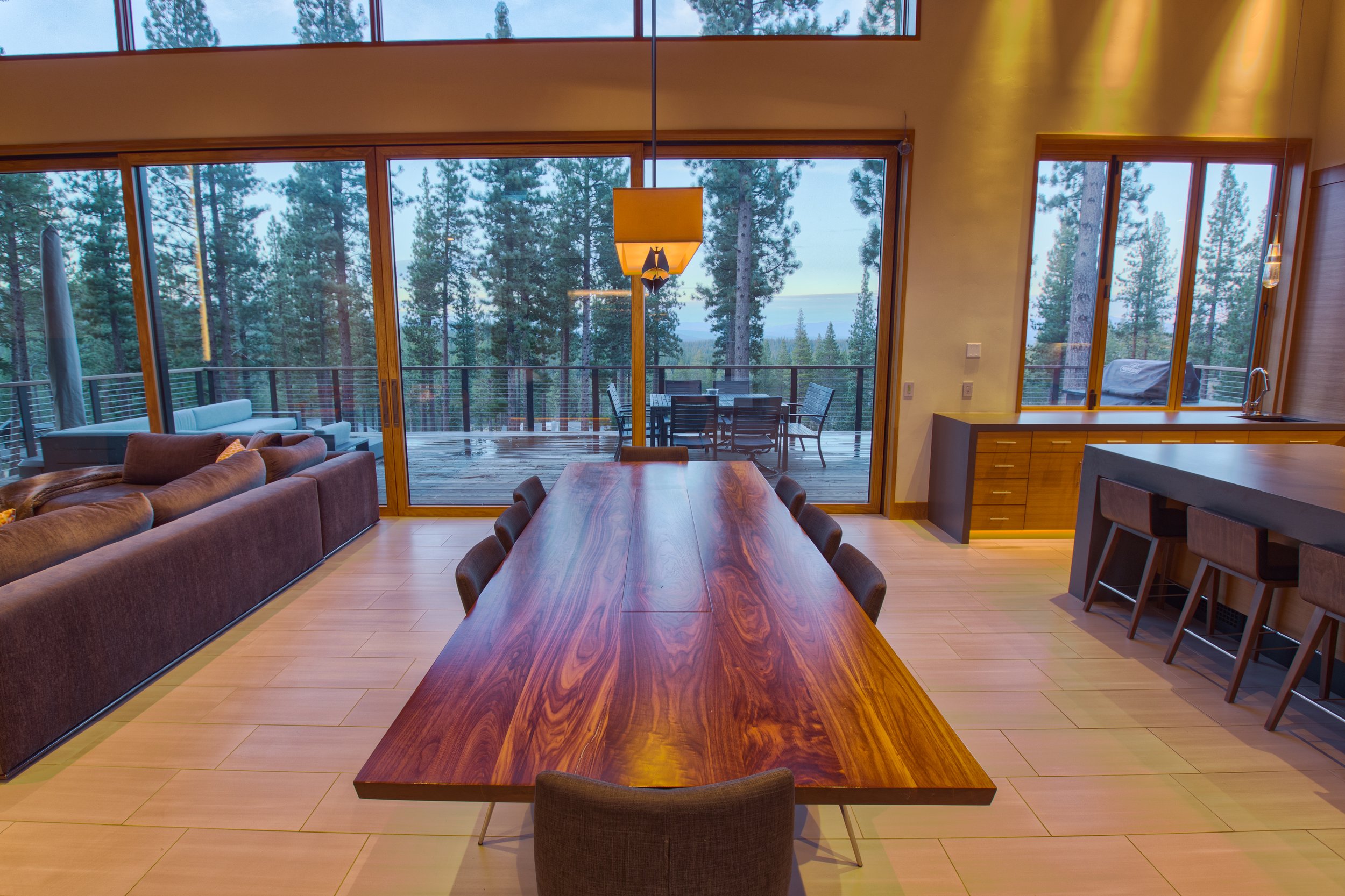 Modern dining room with a long wooden table, six chairs, large floor-to-ceiling windows with a view of a wooden deck and forested landscape, and a nearby kitchen area with bar stools.