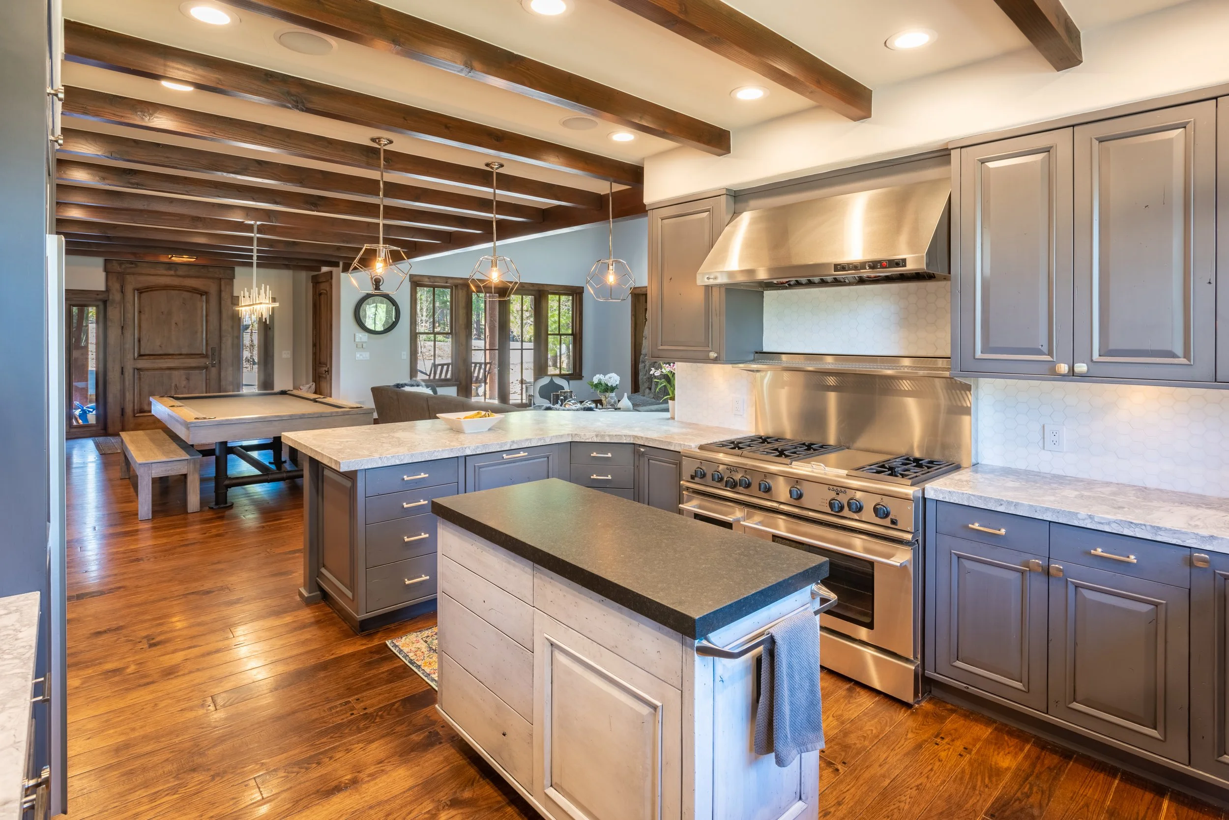 Modern kitchen with gray cabinets, stainless steel appliances, granite countertops, and a wooden beamed ceiling. The space opens to a dining and living area with large windows and hardwood floors.