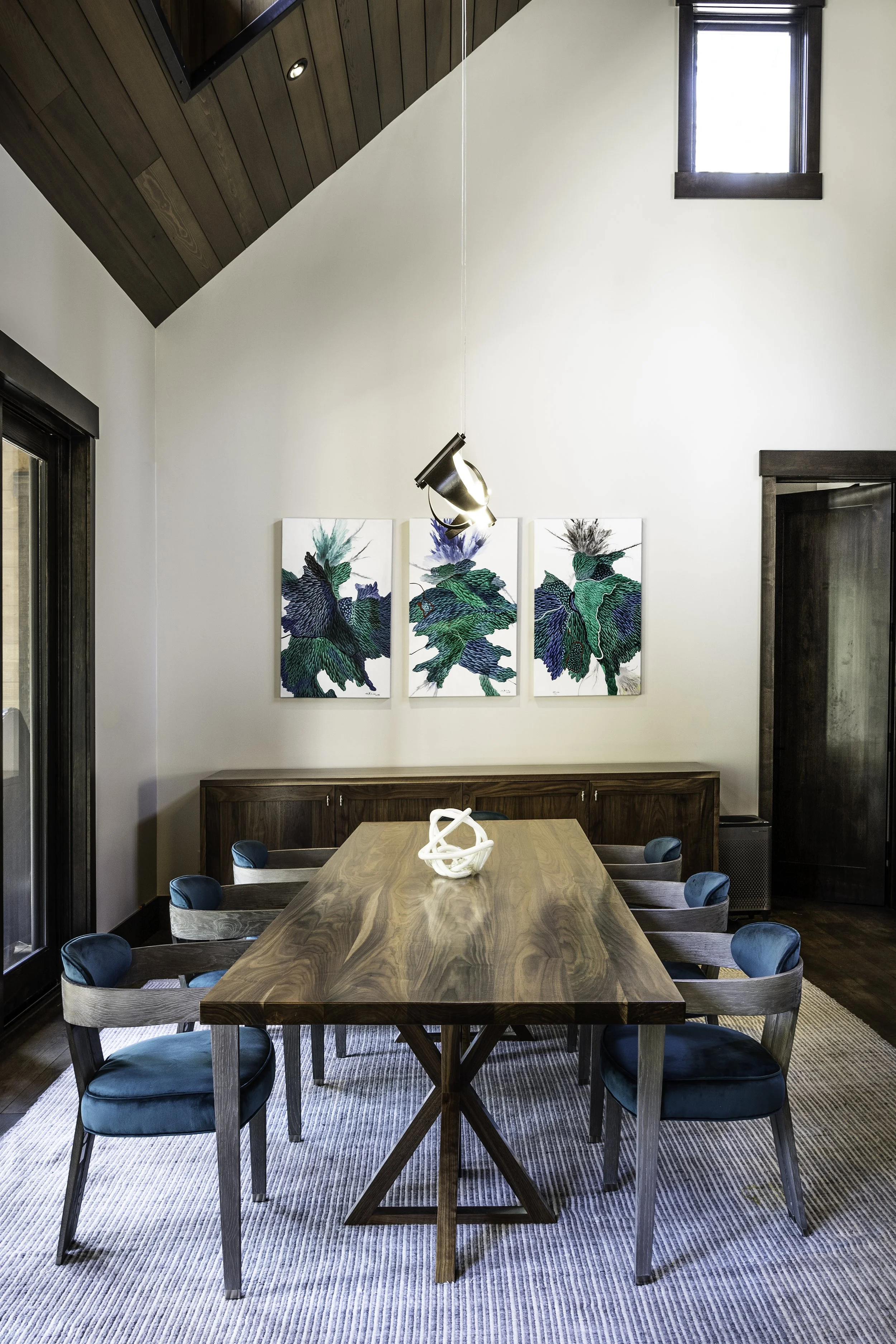 A dining room with an oval wooden table surrounded by eight chairs with teal cushions. There is a striped rug under the table. On the wall, there are three vertical abstract paintings with green and blue hues. A modern white sculpture is on the table