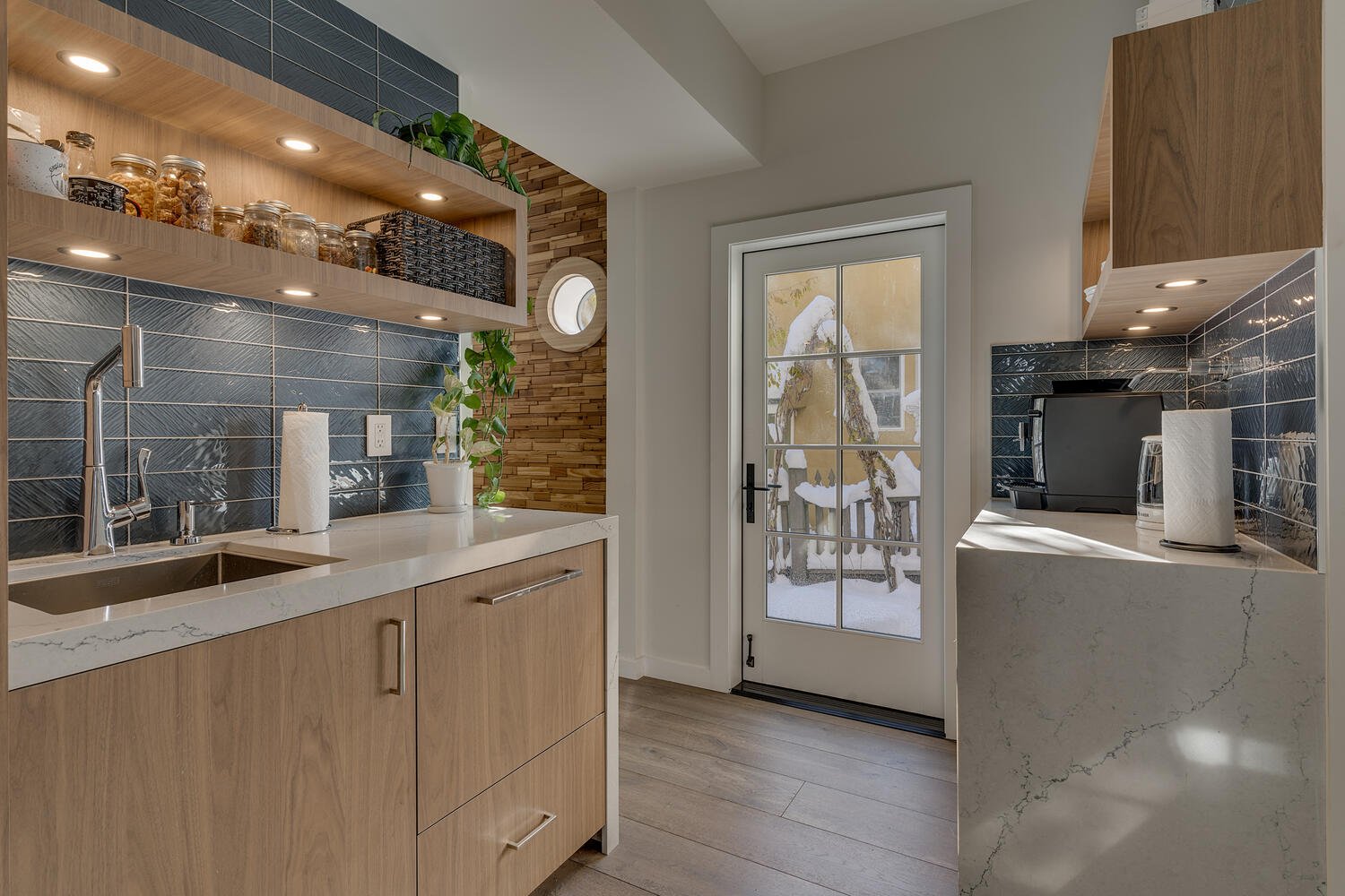 Modern kitchen with wooden cabinets, black tiled backsplash, white countertop, a sink, open shelves holding jars and baskets, potted plants, a door leading outside, and a small counter with a coffee maker and paper towels.