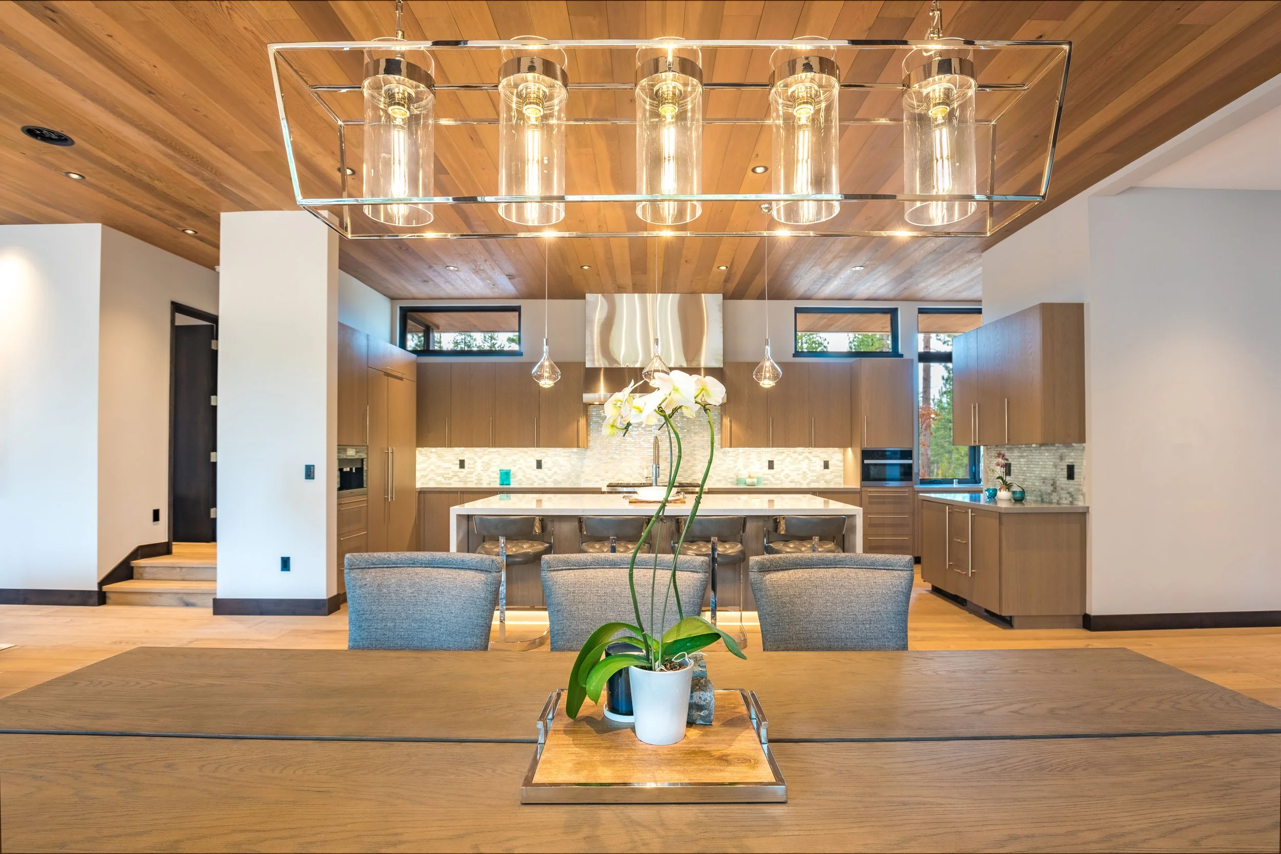 Modern open-concept kitchen with wooden ceiling, pendant lighting, and a central island with bar stools. Dining table with a potted orchid in the foreground.