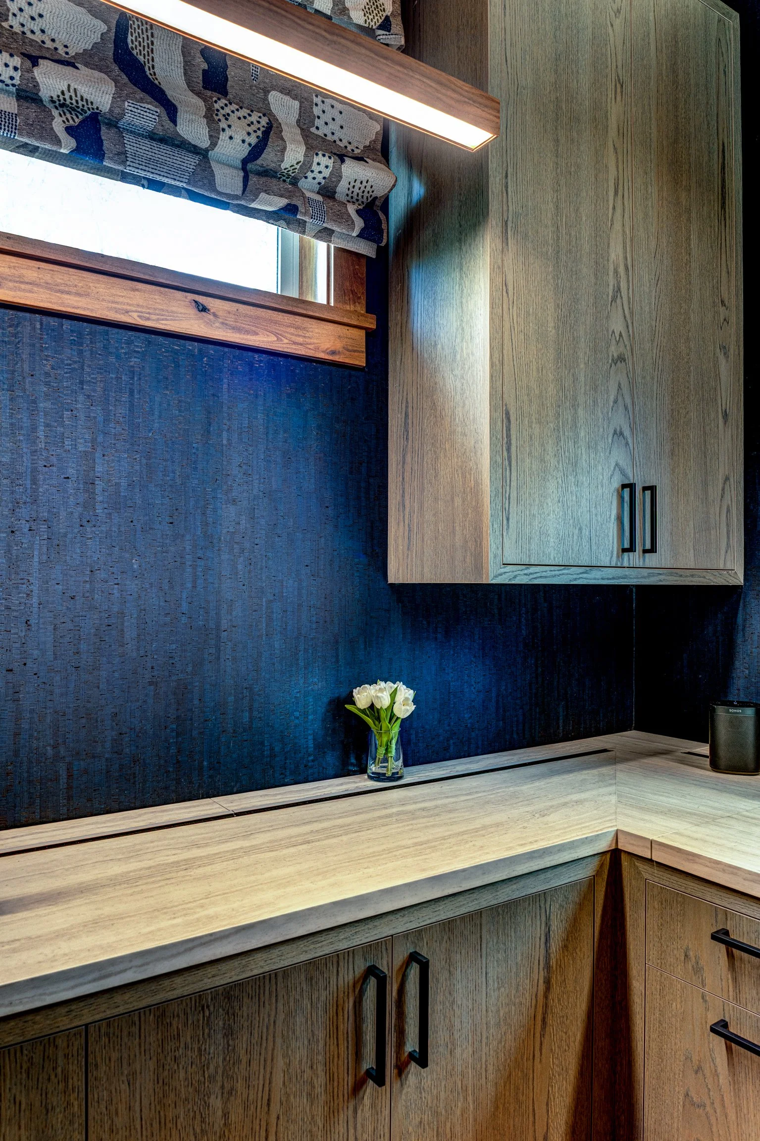 A kitchen corner with wooden cabinets, a blue textured backsplash, a windowsill with a vase of white tulips, and a small black speaker on the countertop.