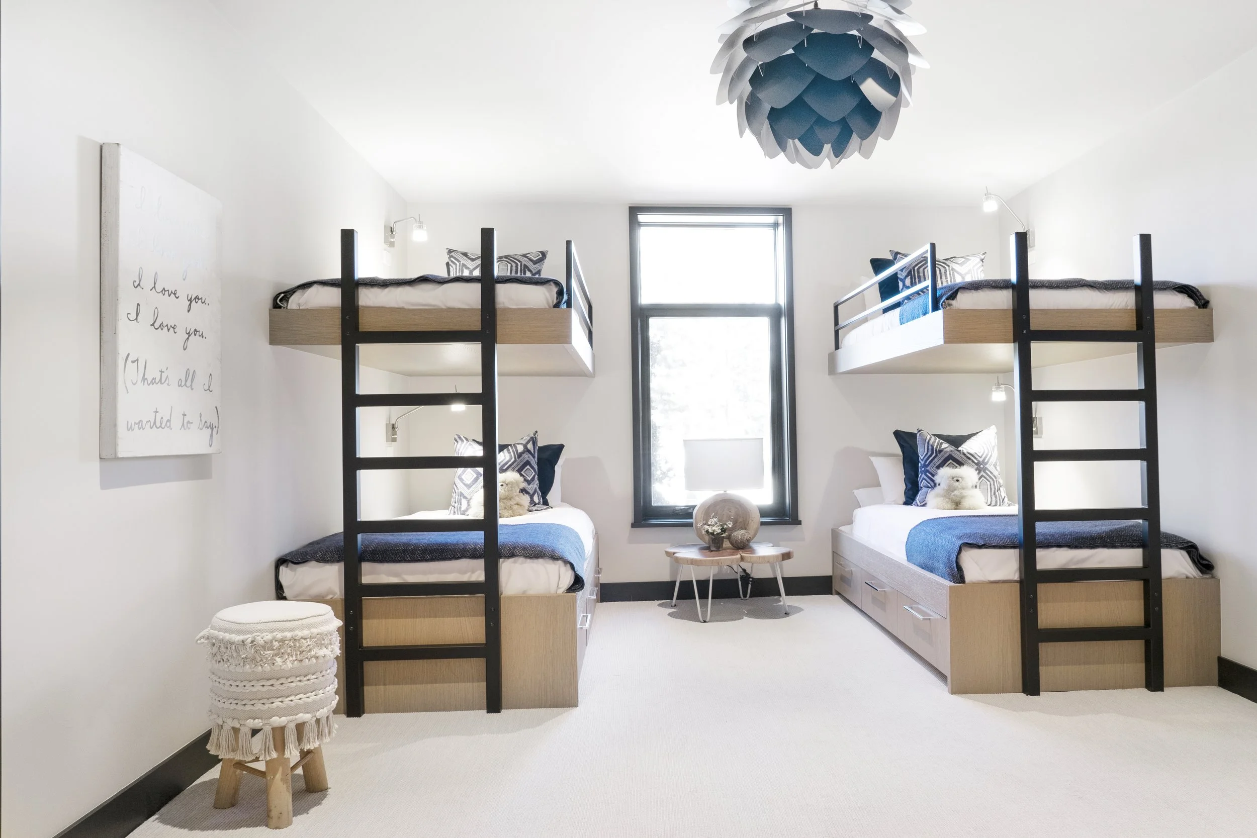 A bedroom with two sets of bunk beds with black ladders against white walls, a window with black trim, a small wooden table with decorative objects in front of the window, a wall art with handwritten notes on the left, and a round ottoman in the fore