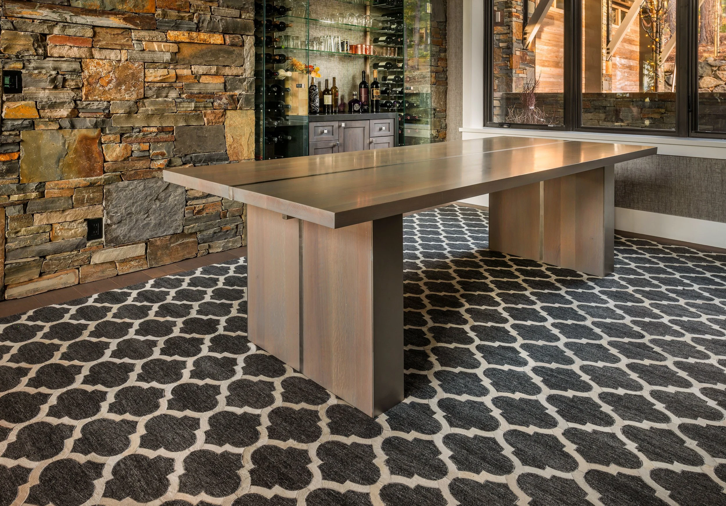 Modern dining room with a large wooden table, a stone accent wall, a wine display, and large windows with a view of trees outside.