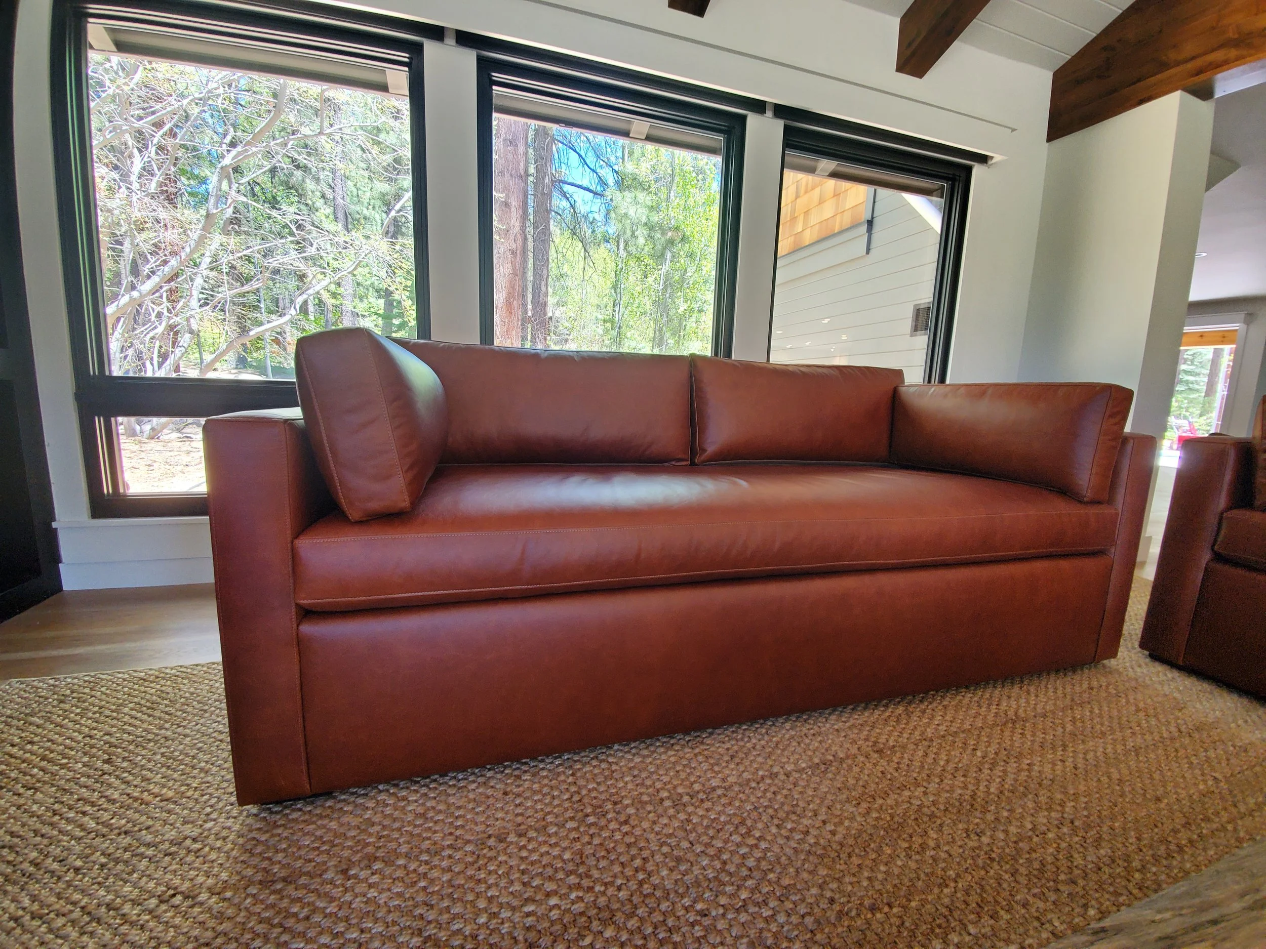 A brown leather sofa with square armrests, placed on a woven rug in front of large windows showing trees outside.