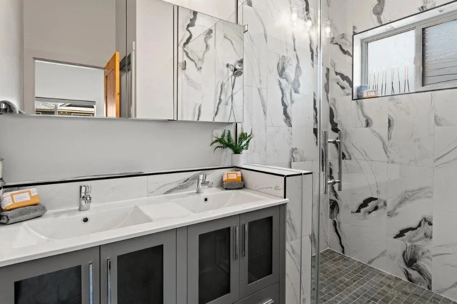 Modern bathroom with marble walls, double sink vanity, gray cabinets, and glass-enclosed shower with a window.