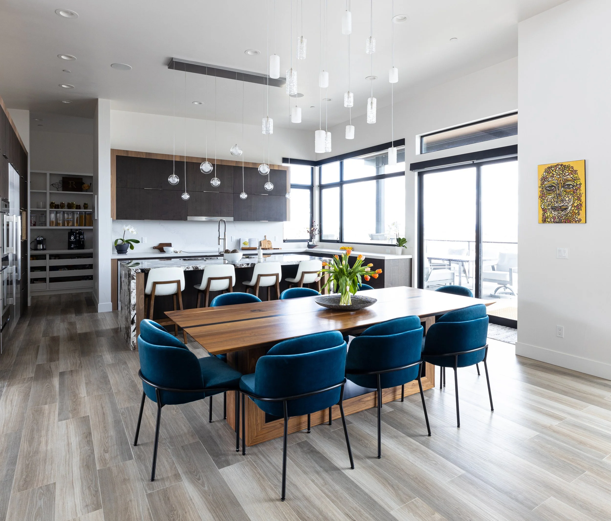 Modern open-concept kitchen and dining area with wooden table, blue velvet chairs, a kitchen island with white and dark cabinetry, large windows, and a colorful artwork on the wall.
