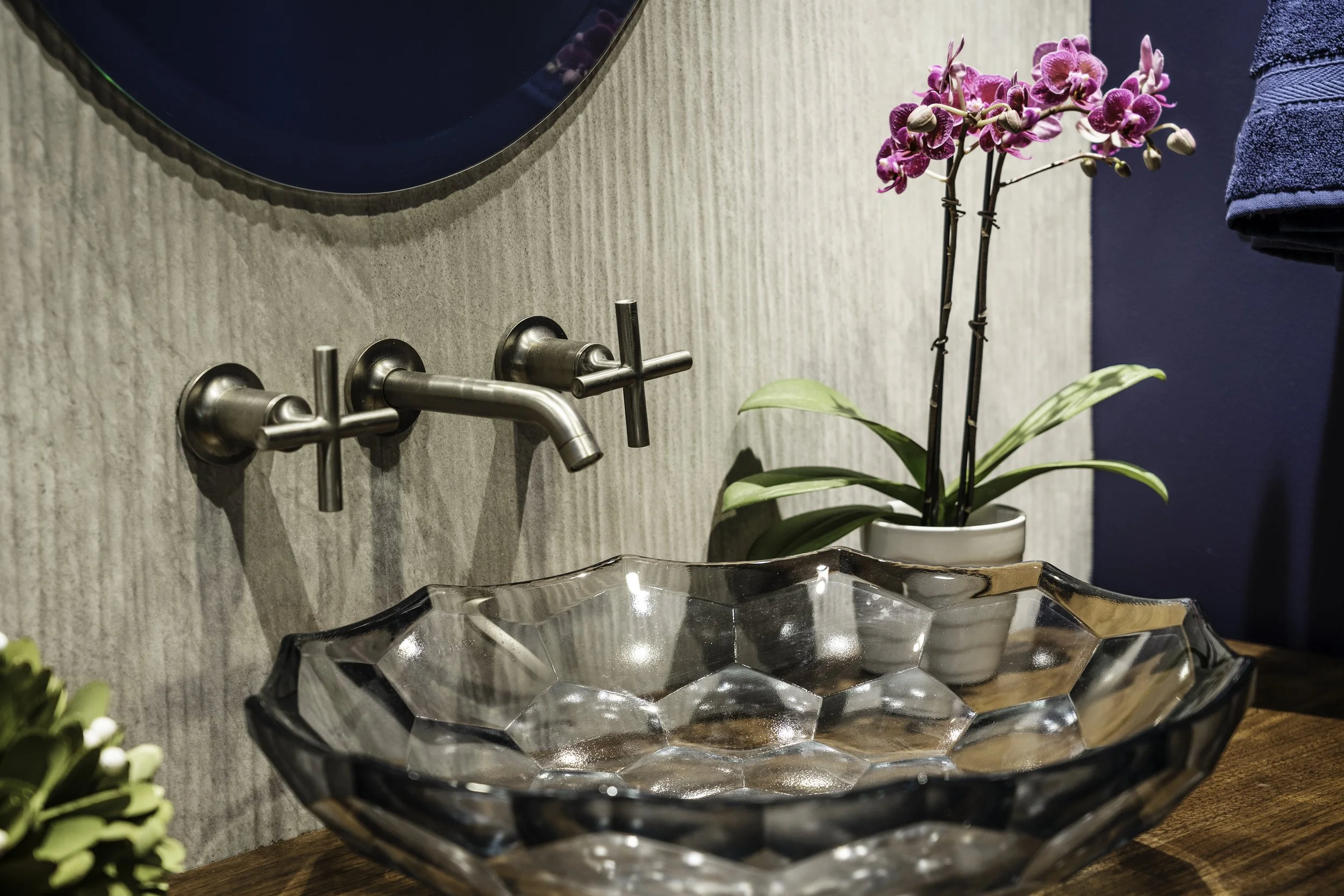 A glass sink with angled edges on a wooden countertop, a wall-mounted faucet above it, and a potted orchid flower plant beside it.