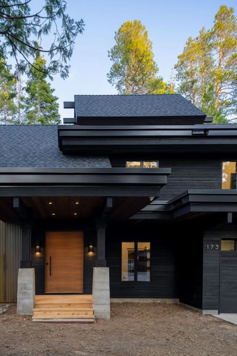 Modern black house with wooden front door, surrounded by tall trees, with a small front porch and steps leading to the entrance.