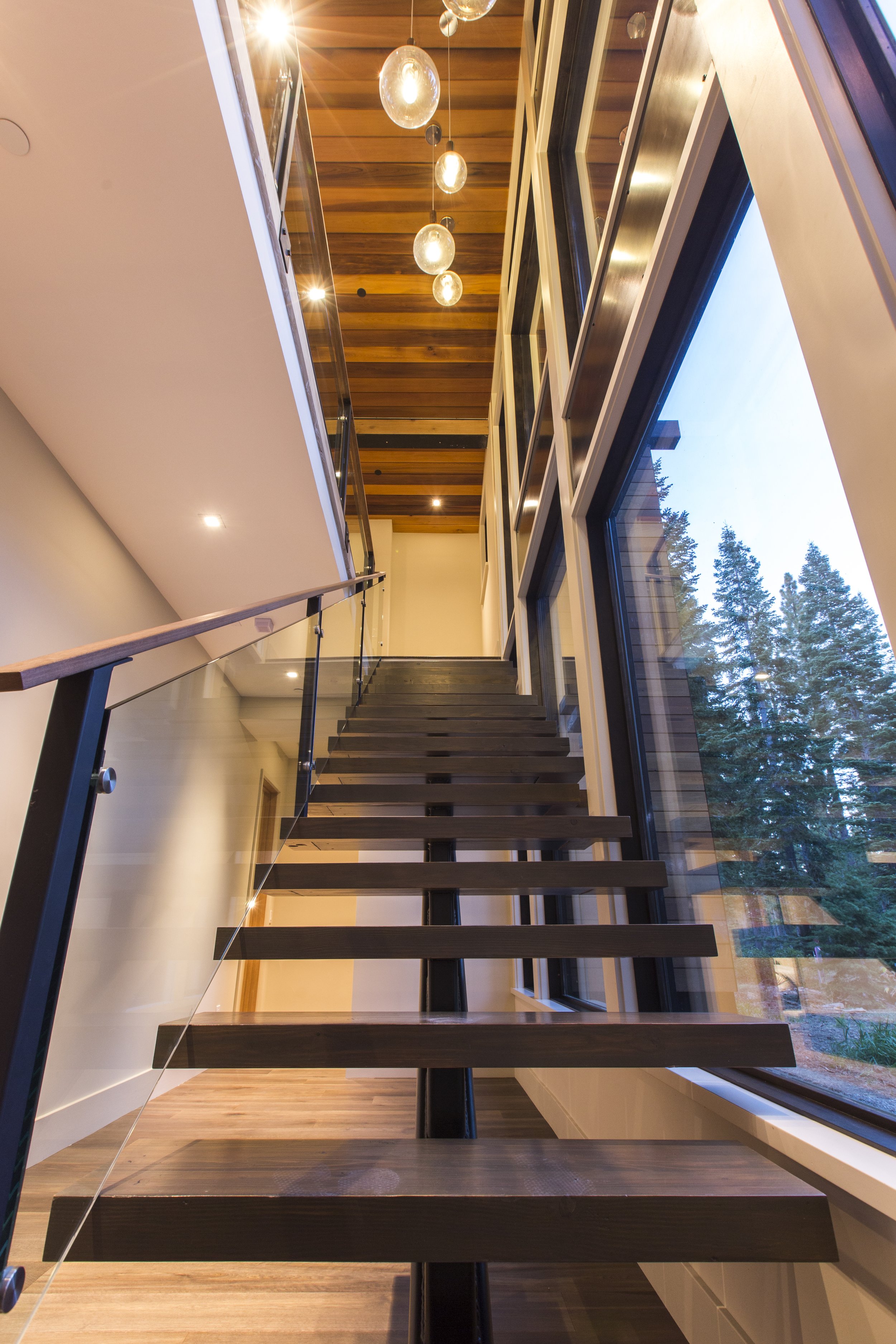 Interior view of a modern staircase with dark wooden steps, glass railing, and large windows overlooking trees, with pendant lights hanging from a wooden ceiling.