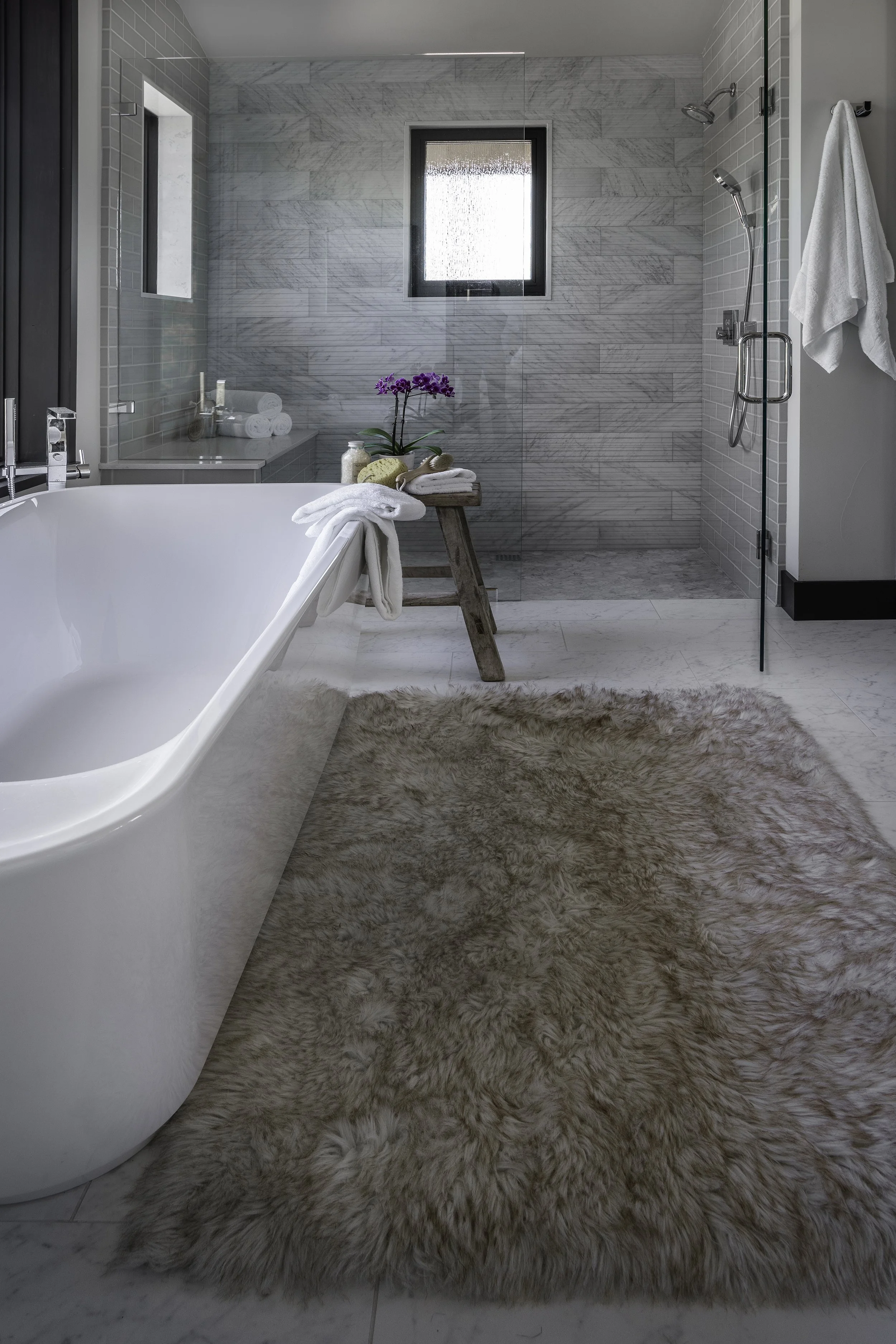A modern bathroom with a freestanding bathtub, a fluffy beige rug, and a walk-in shower with gray marble tiles and a window, decorated with a purple orchid on a wooden stool.