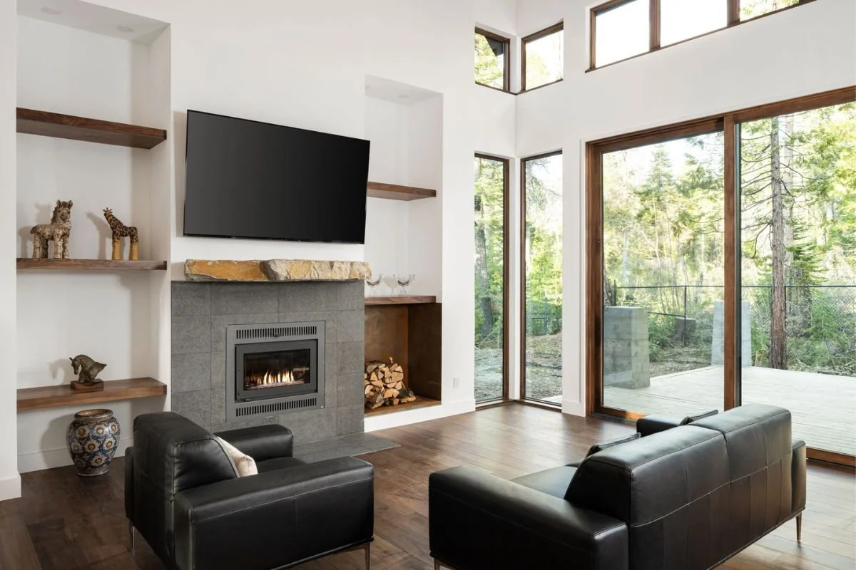 Modern living room with a fireplace, flat-screen TV, large windows with green outdoor view, black leather sofa, and wooden shelves with decorative items.