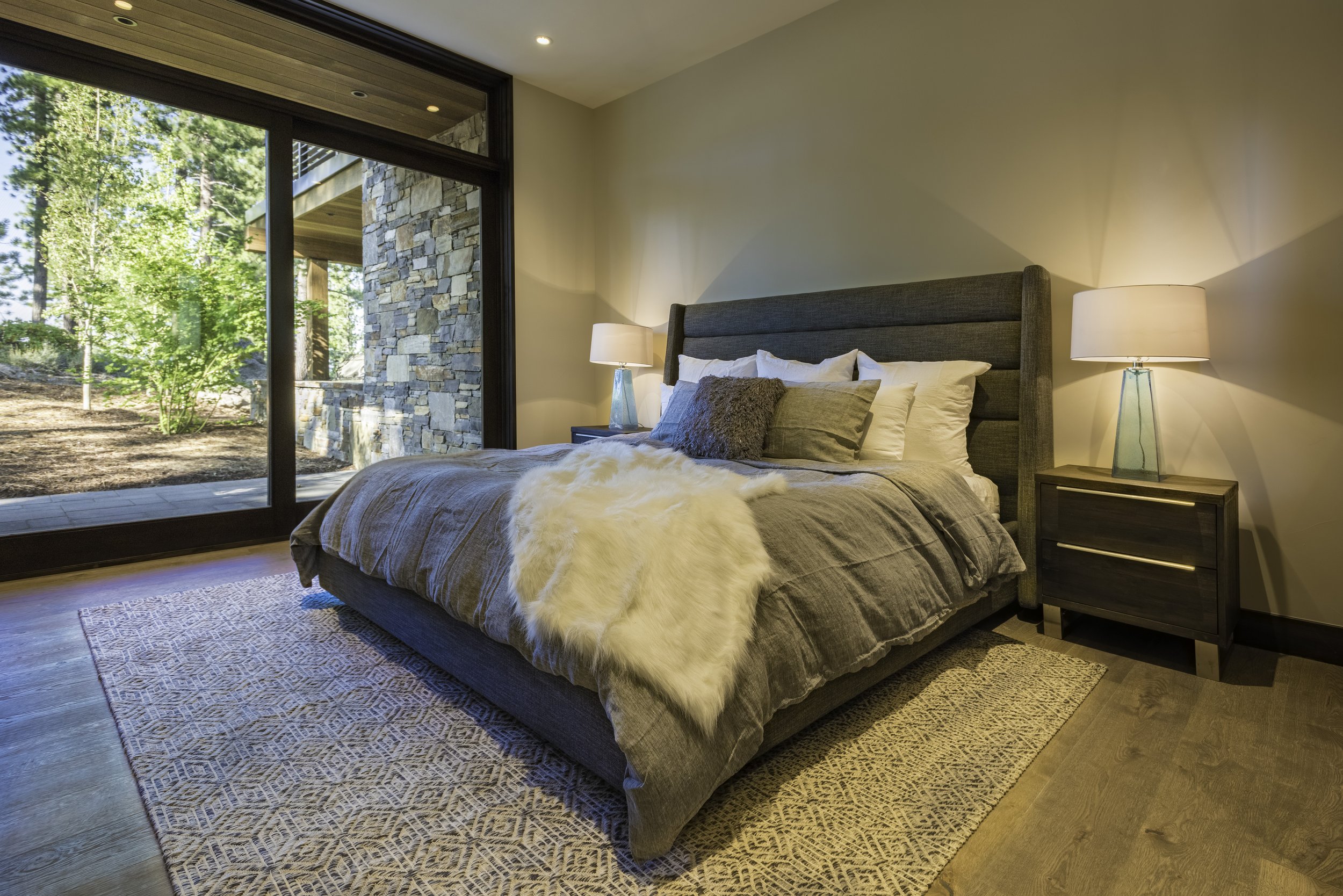 Modern bedroom with large bed, gray upholstered headboard, beige bedding, and decorative pillows, illuminated by two lamps on black nightstands, with sliding glass door revealing an outdoor area with trees and stone wall.