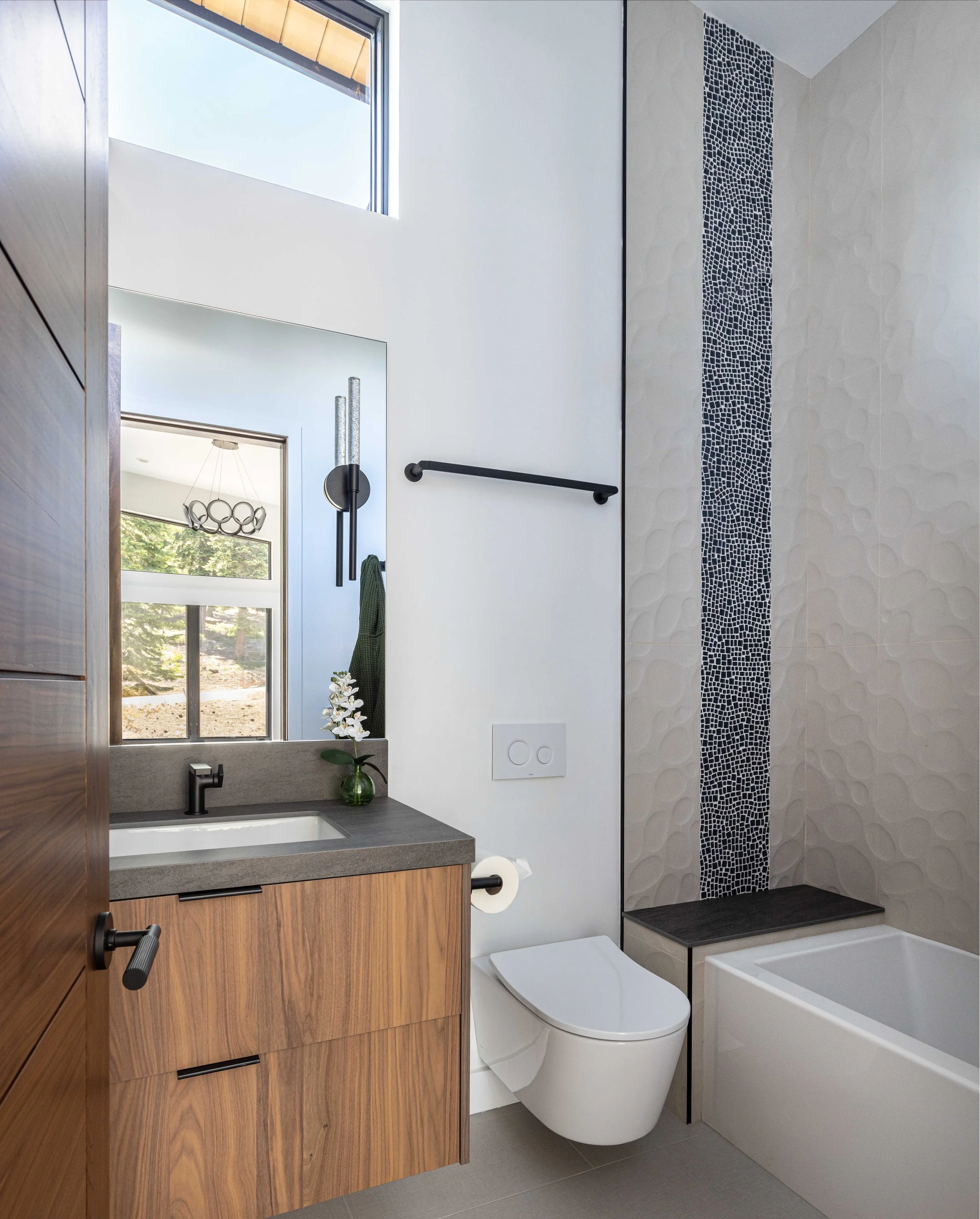 Modern bathroom with a wooden vanity, countertop sink, potted plant, wall mirror, black wall-mounted bathroom accessories, a toilet, and a bathtub with a textured, stone-like wall and a narrow black mosaic strip.