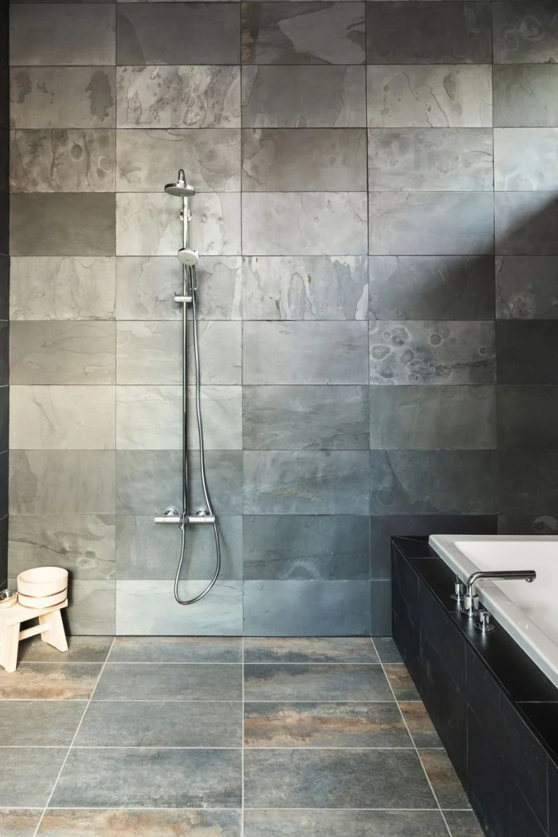 Modern bathroom with gray stone tile walls and floor, a showerhead and hose on the wall, and a black bathtub to the right, with a small wooden stool holding rolled towels in the corner.