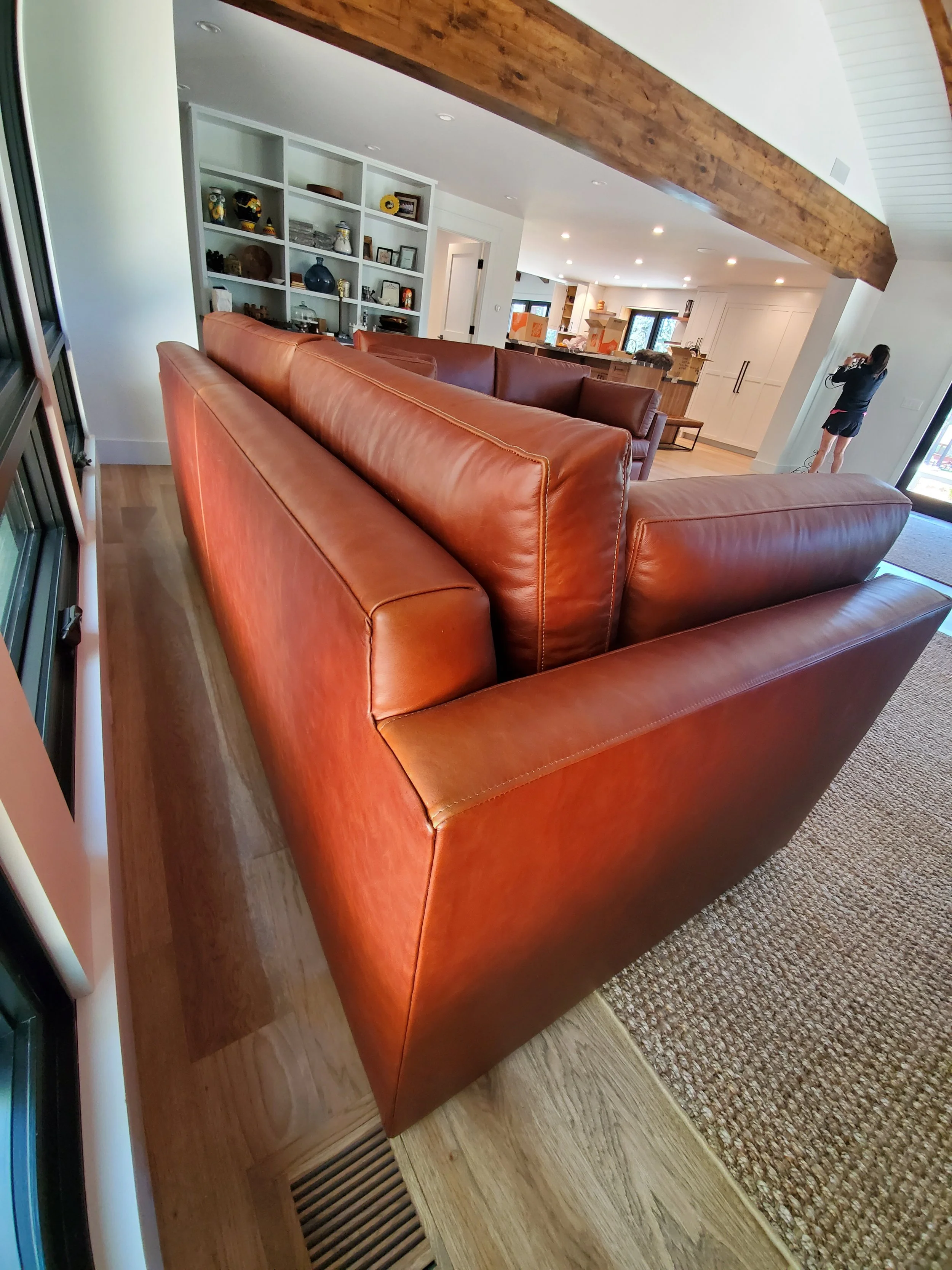 Living room with a large brown leather sofa, a white built-in bookshelf with decorative items, a wooden beam on the ceiling, hardwood floors, and a person taking a photo near a sliding glass door.
