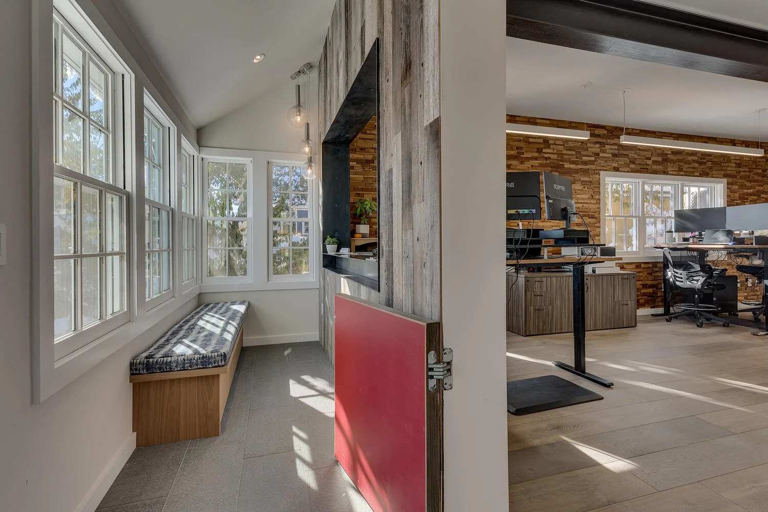 Bright sunlit corner with a cushioned window seat, multiple large white-framed windows, and a small indoor plant, adjacent to a modern open office area with wooden accent wall, desks, and chairs.