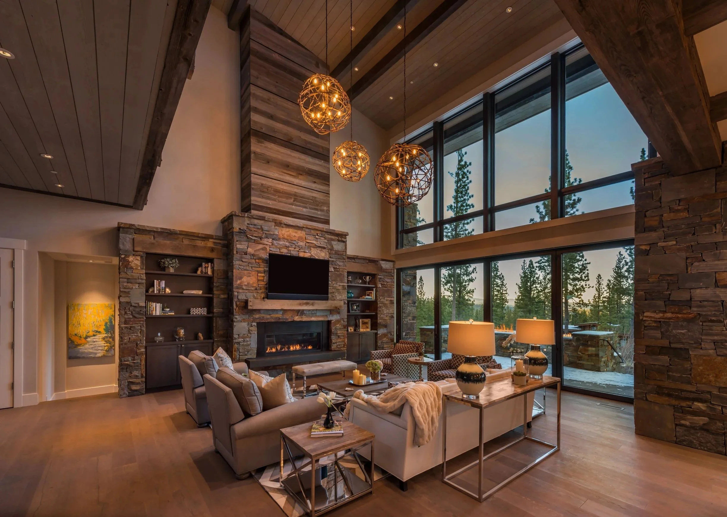 Living room with high ceiling, large windows showing trees outside, a stone fireplace, and modern furniture including sofas, armchairs, and lamps.